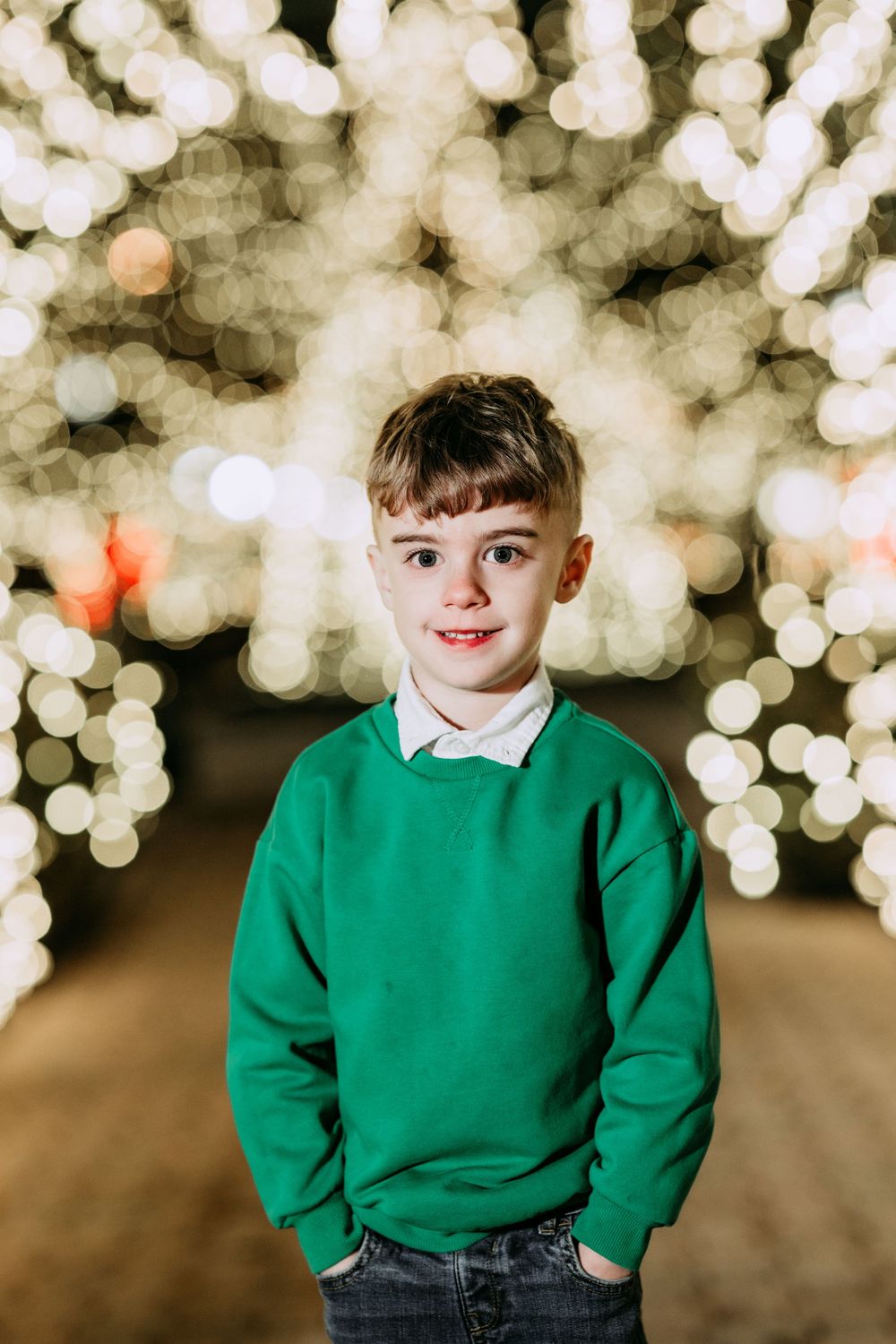 Child in green school sweater smiles against blurred bokeh lights background creating festive holiday atmosphere.