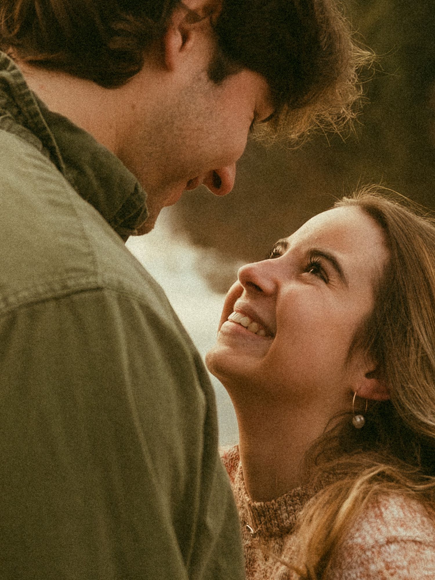 A smiling woman gazes up at a man in a warm, romantic moment, both sharing an intimate connection outdoors.