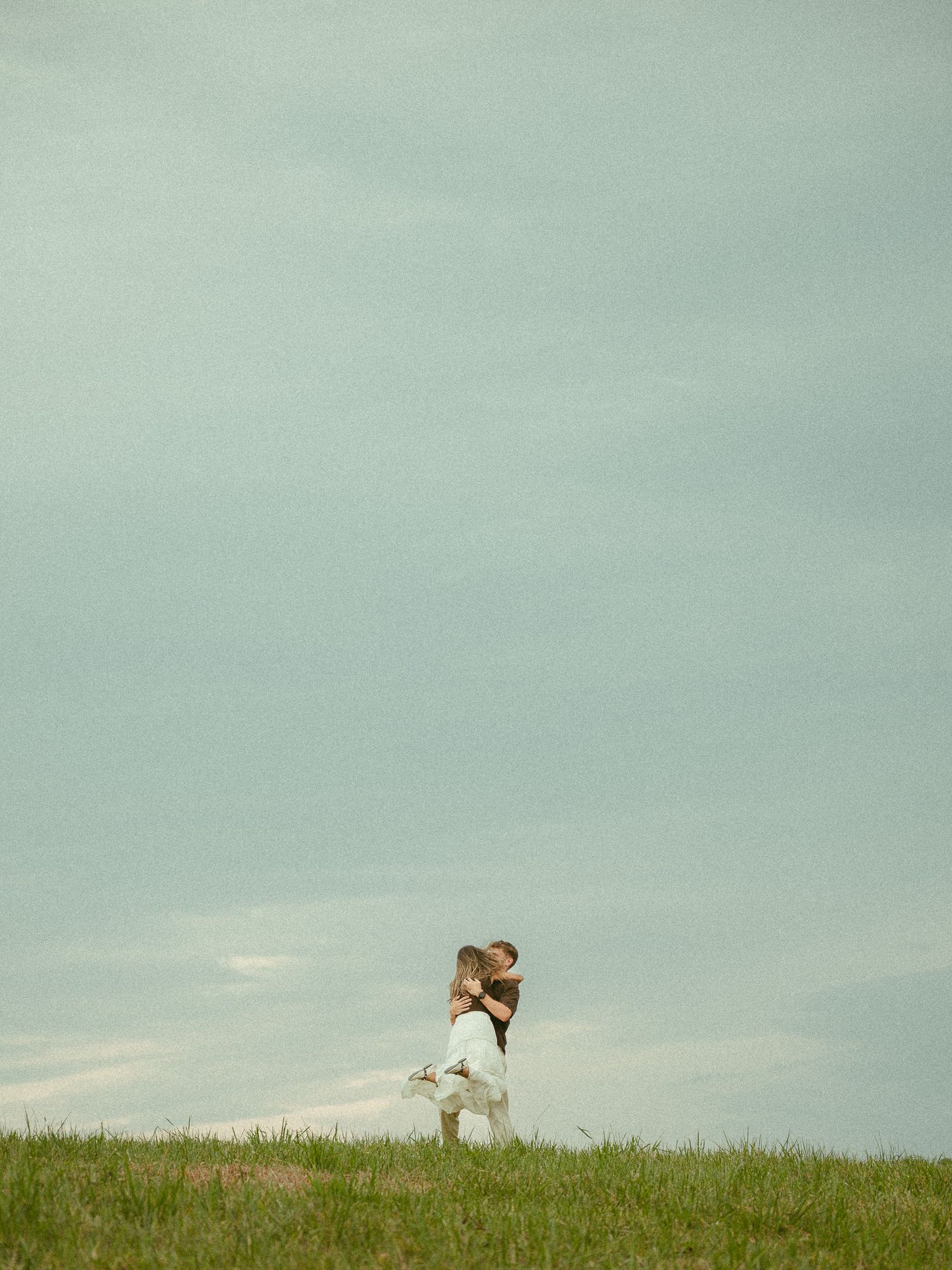 A couple embraces romantically on a grassy hill beneath a dramatic cloudy sky in an open field.