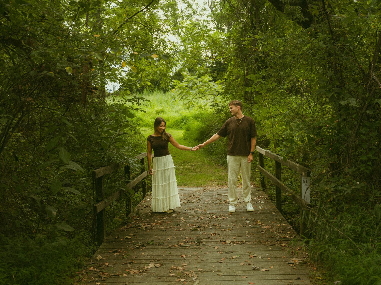 Couple holding hands on a wooden bridge surrounded by lush green trees in a forest.
