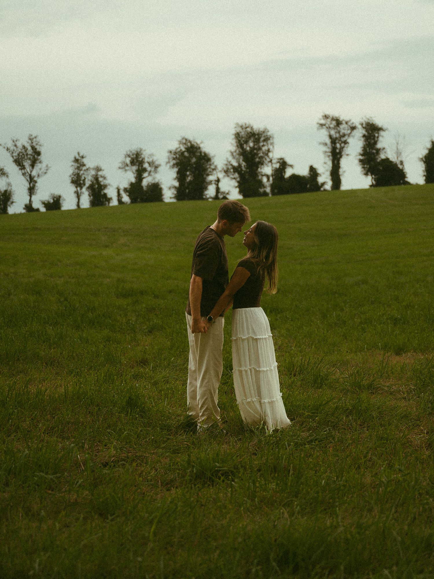 Romantic couple standing close in a lush green field, about to kiss, with trees on the horizon.