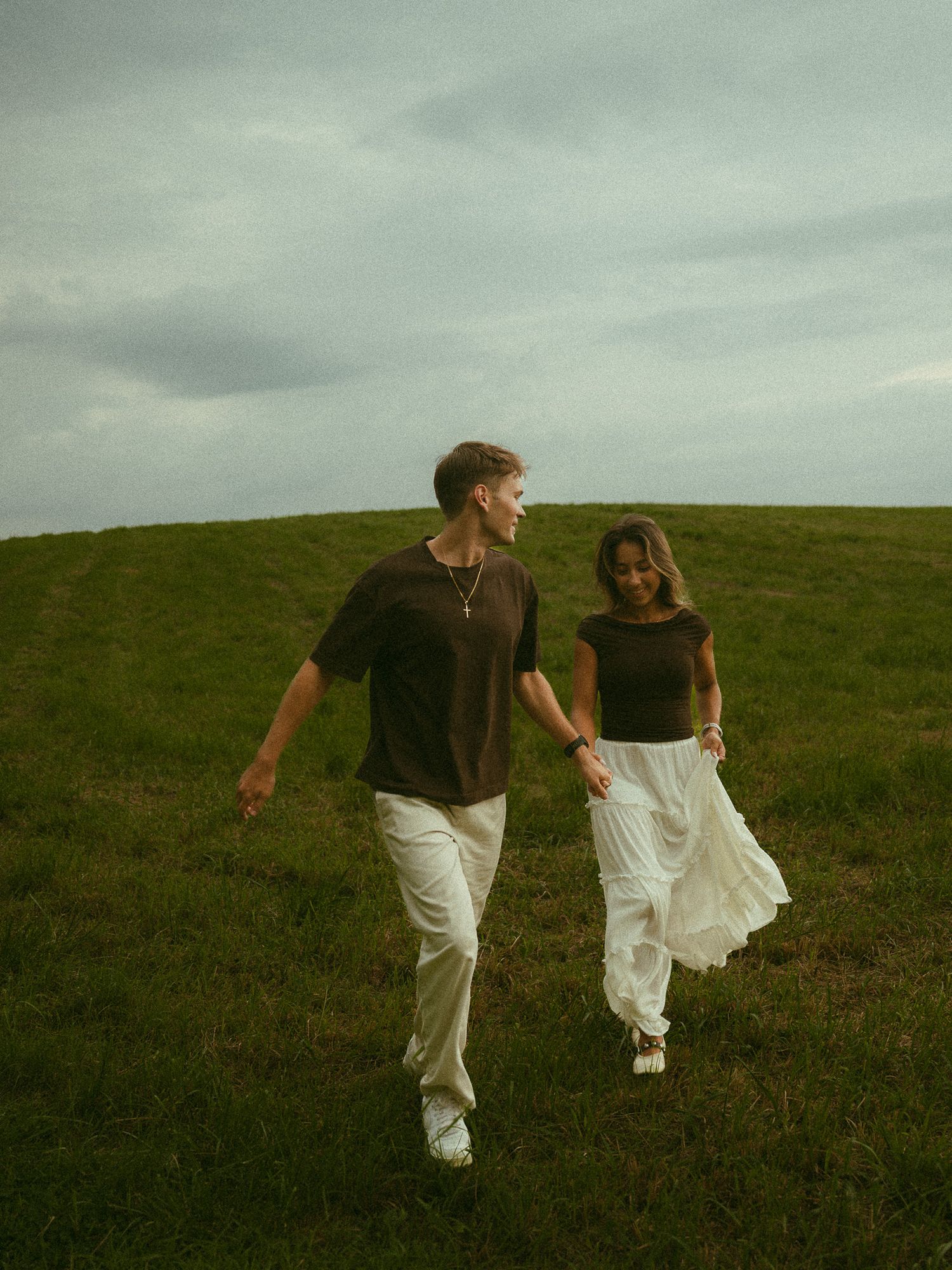 Couple walking hand in hand through a green grassy field under a cloudy sky.