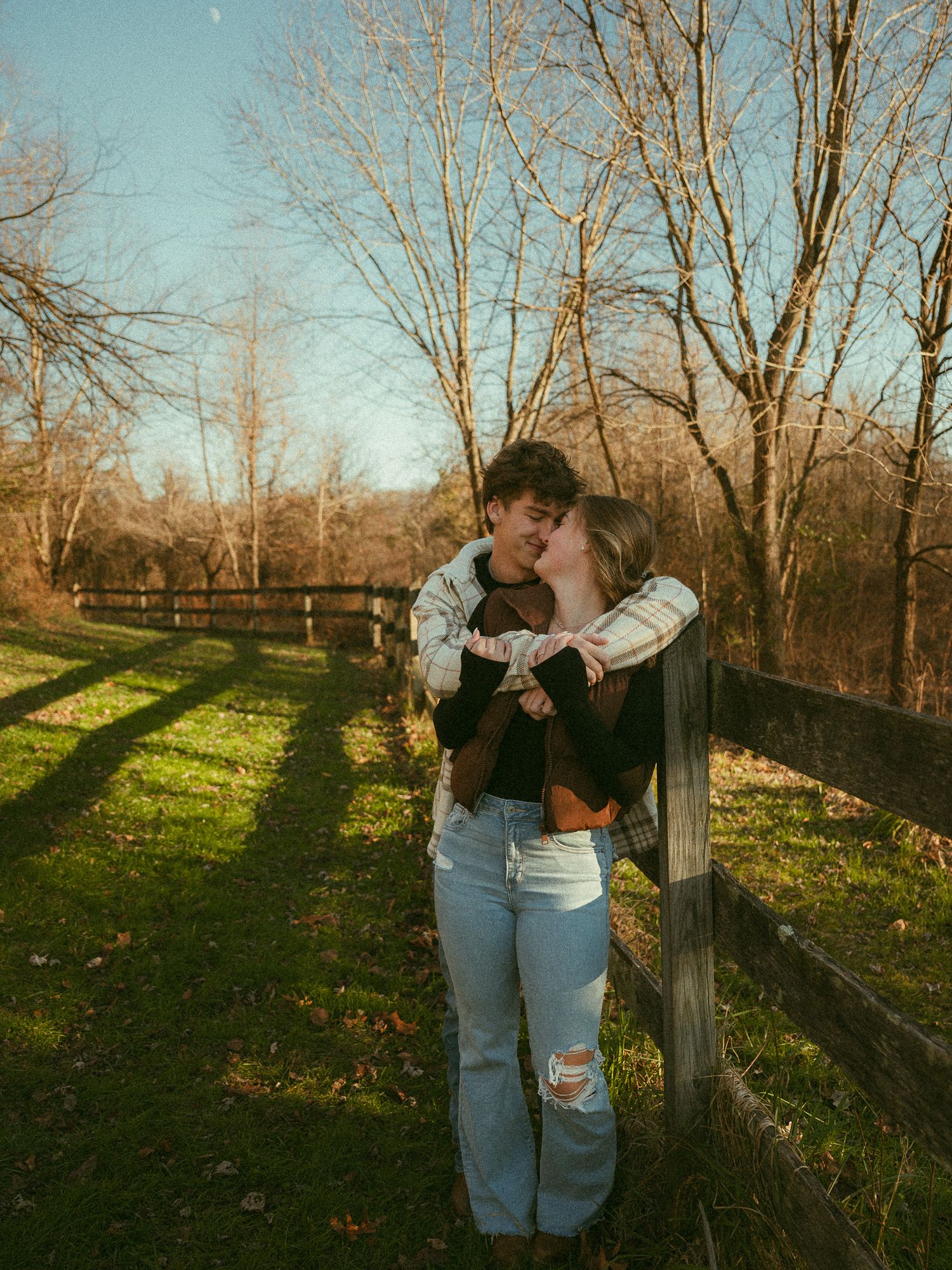 Couple embracing and kissing by a wooden fence in a sunny autumn field with bare trees.