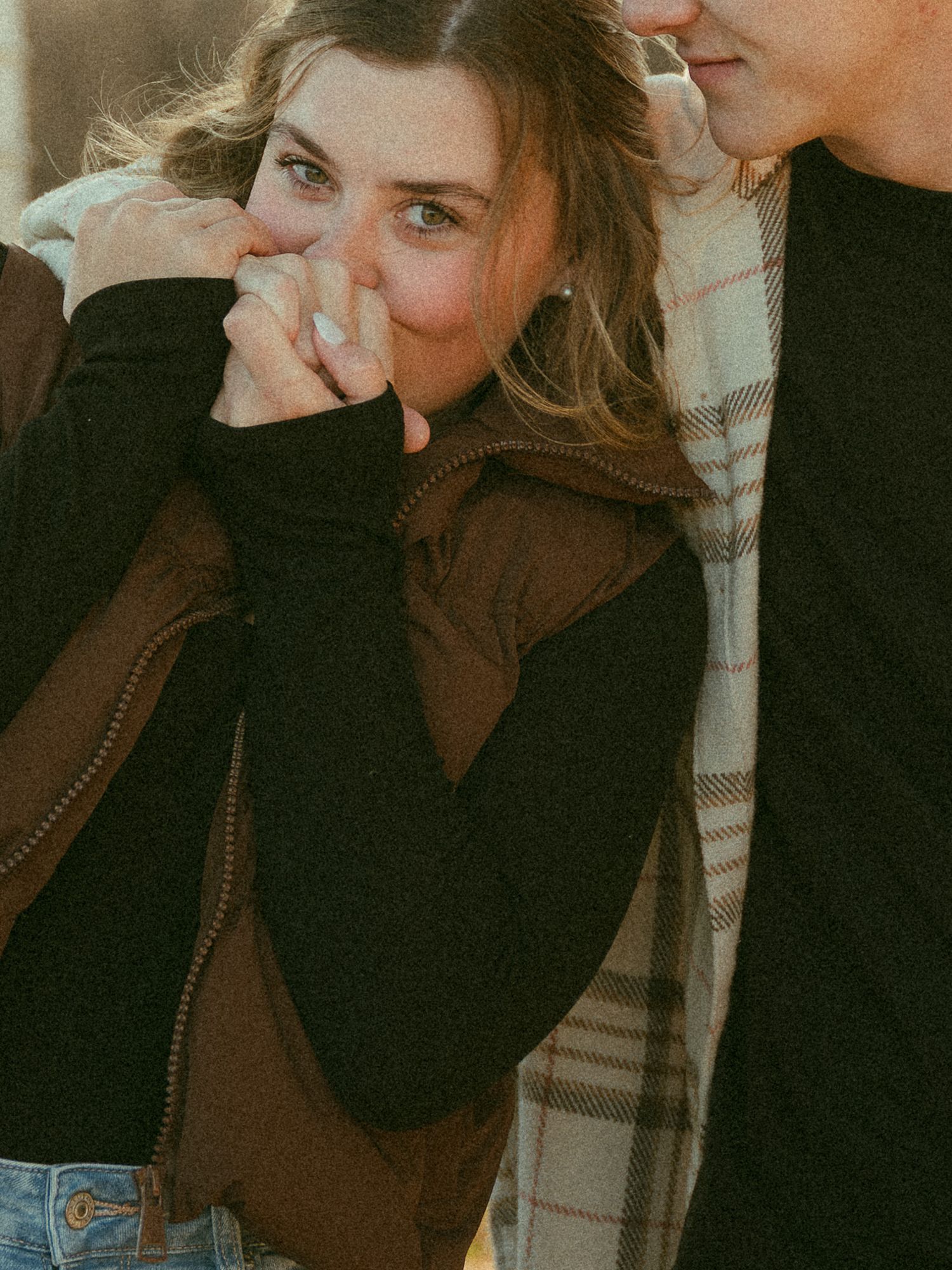 A woman laughing shyly, covering her mouth, wearing a brown vest and black jacket outdoors.