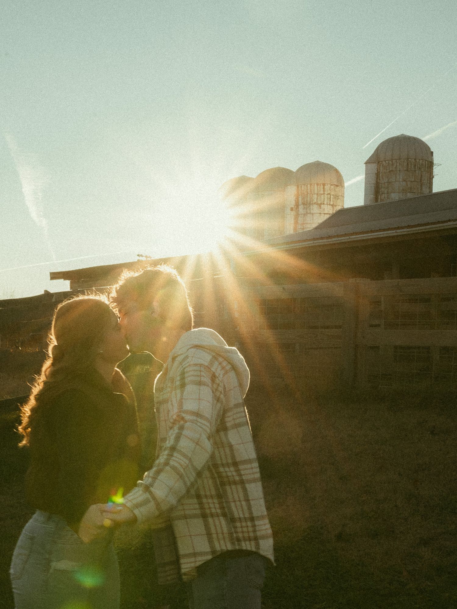 Couple kissing at sunset with farm silos silhouetted against bright sunburst in background.