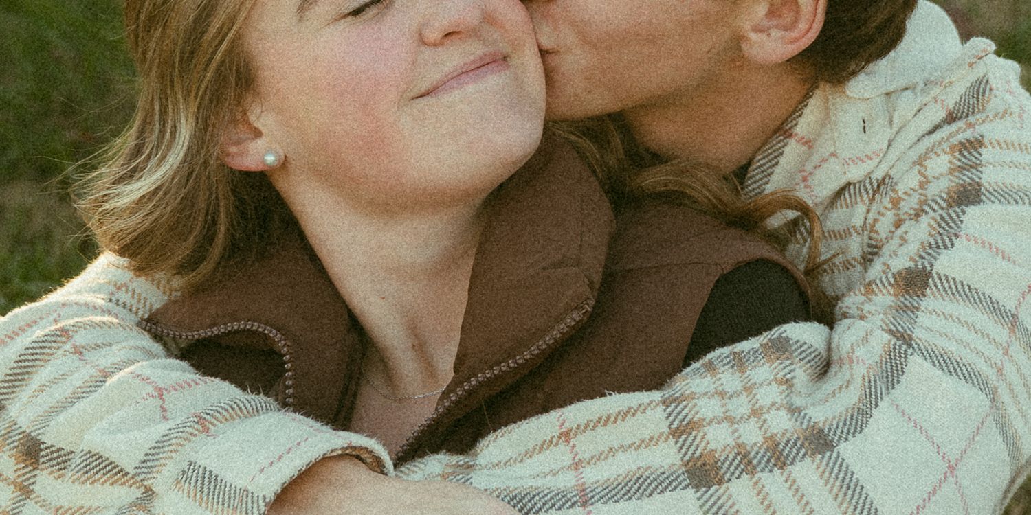A couple sharing a tender kiss and embrace outdoors, woman smiling in brown vest and plaid shirt.