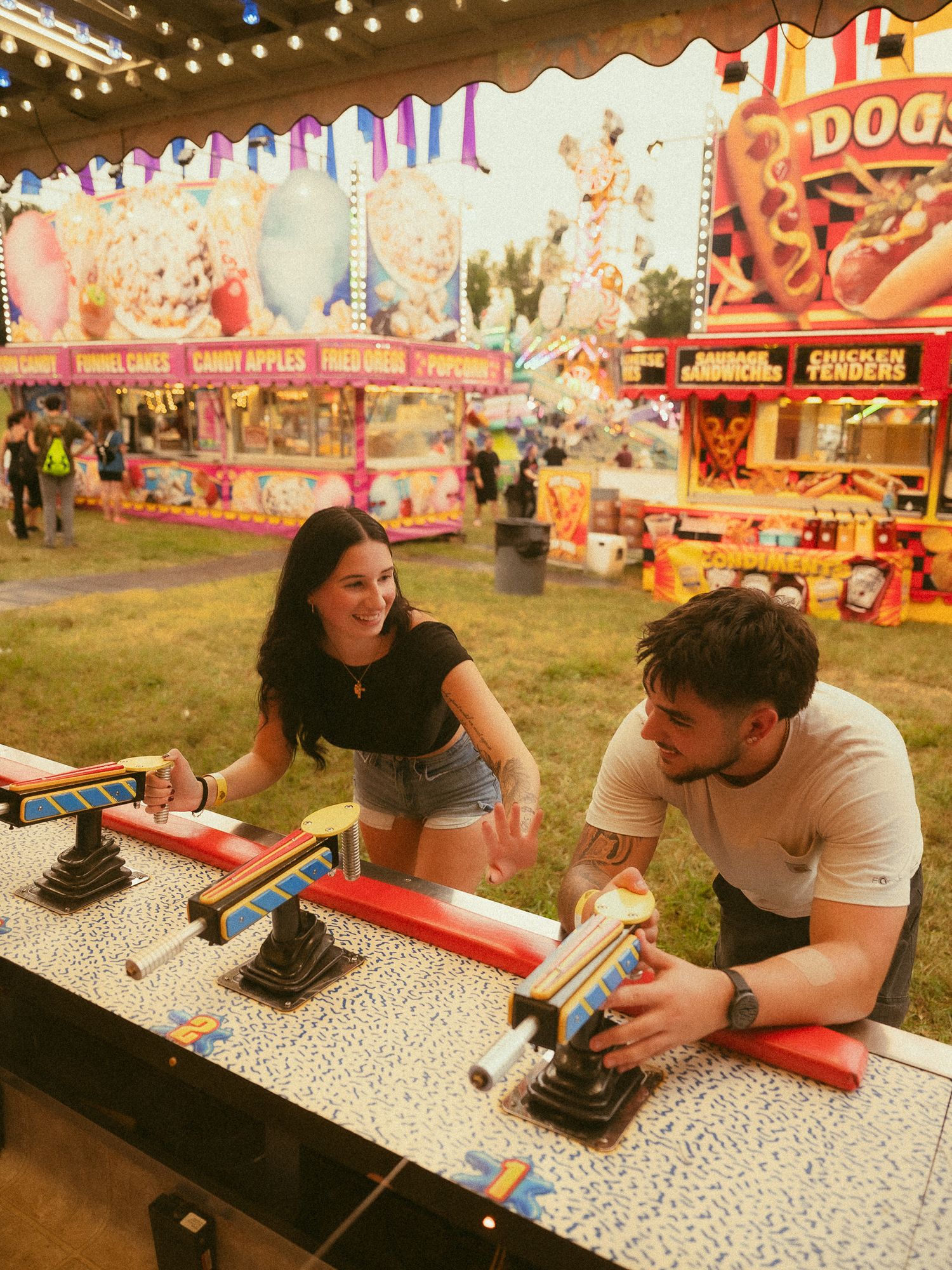 A couple plays a carnival game at a colorful fairground with food stands selling hot dogs and chicken tenders in the background.