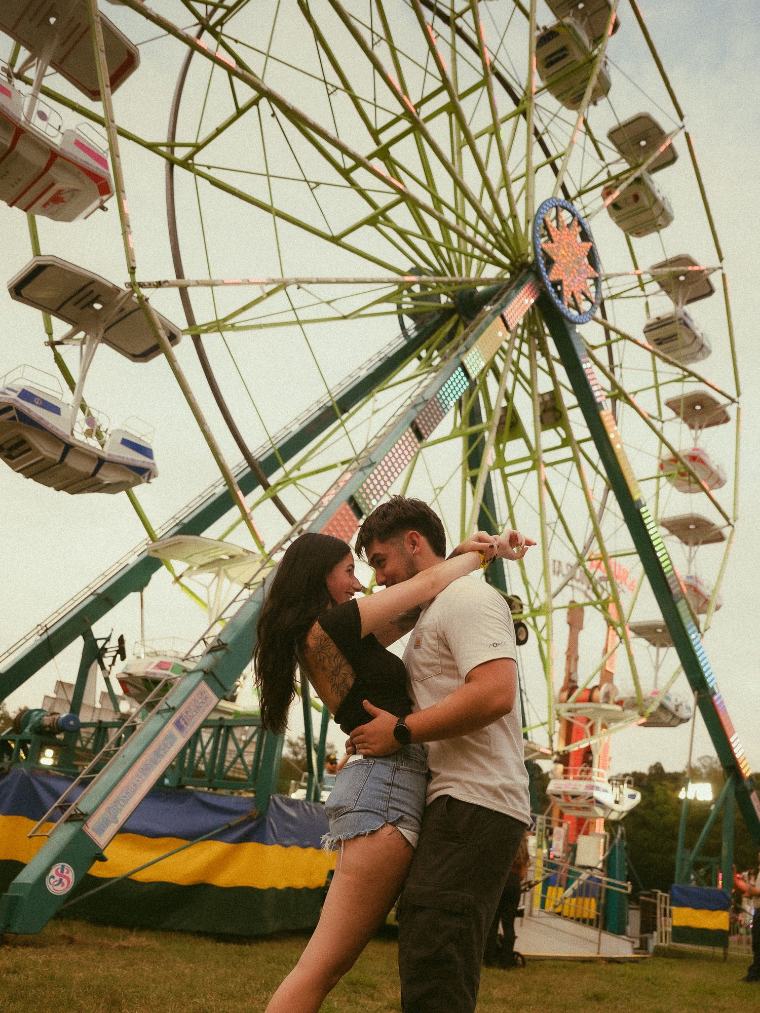 Couple embracing romantically in front of a colorful ferris wheel at a summer carnival fair.