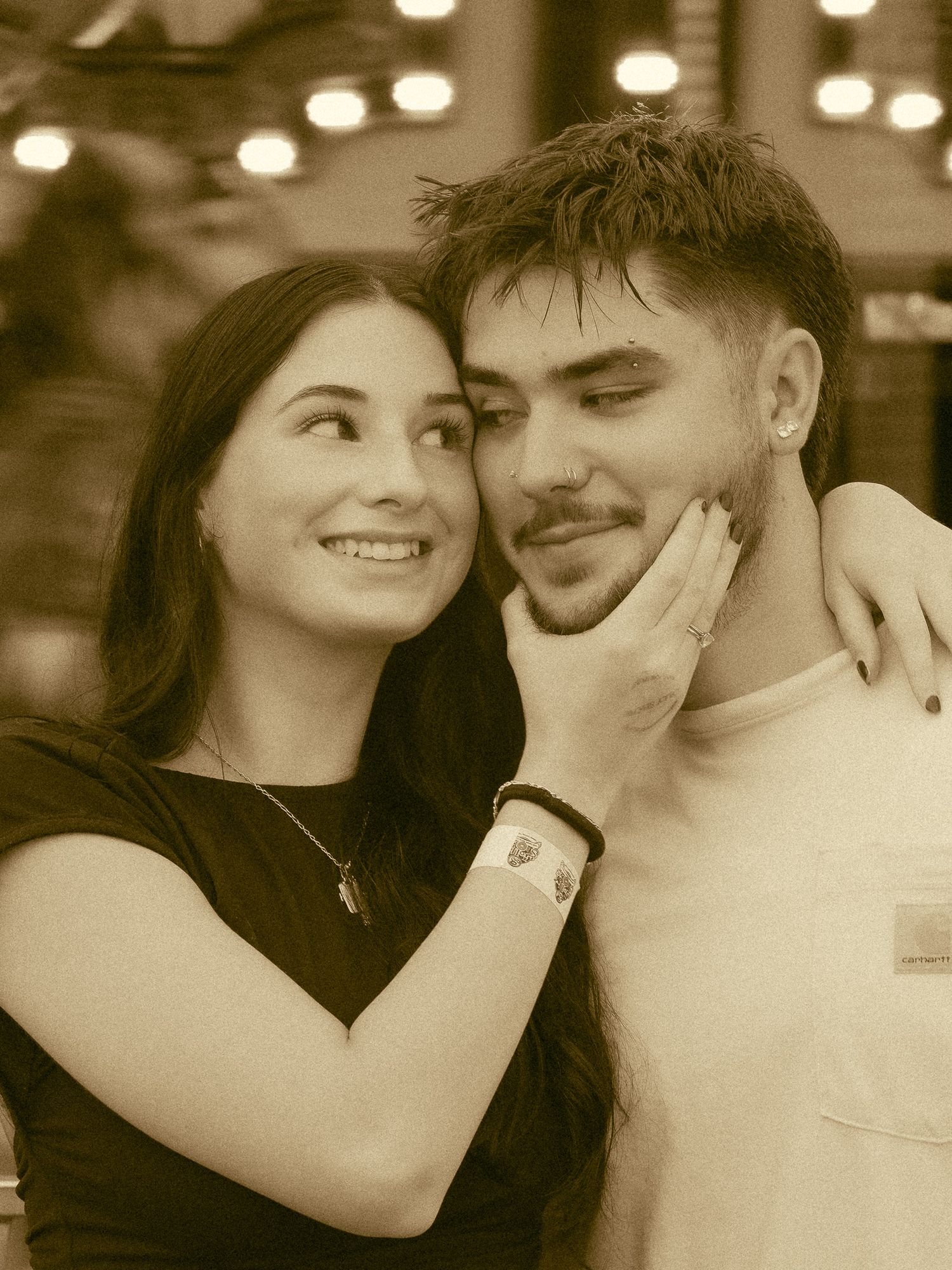 A young couple shares a tender moment, with the woman smiling and gently touching the man's face in a warm sepia-toned photo.