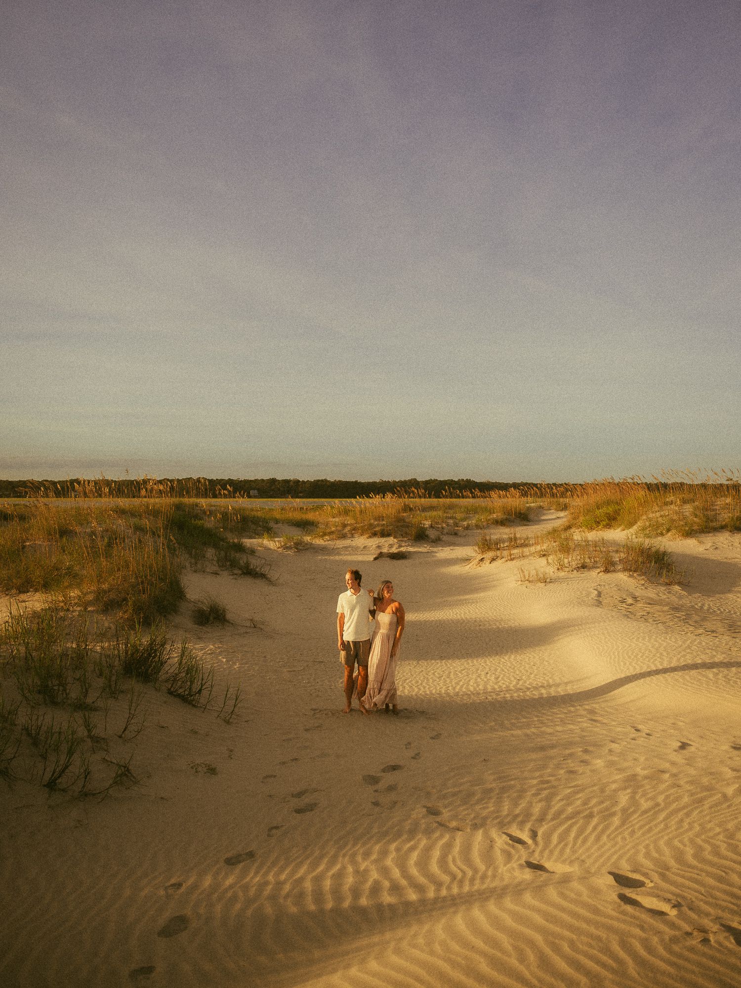 Couple standing together on sandy beach dunes at golden hour sunset with grass and trees in background.