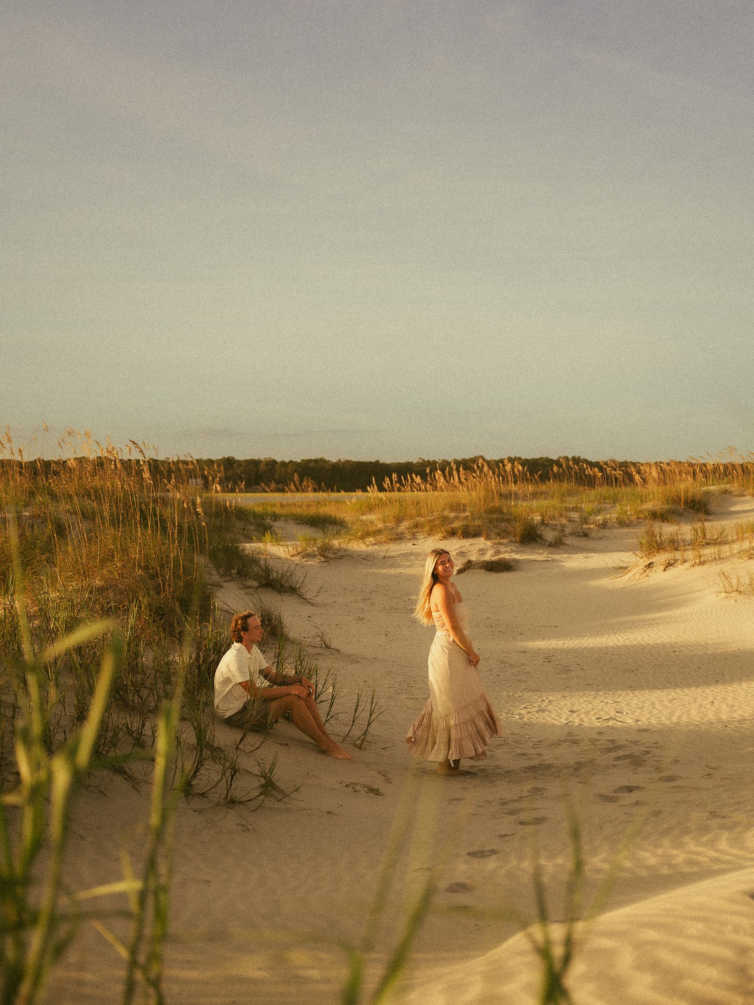A couple on sandy beach dunes at golden hour, woman in flowing dress standing near man seated among sea oats.