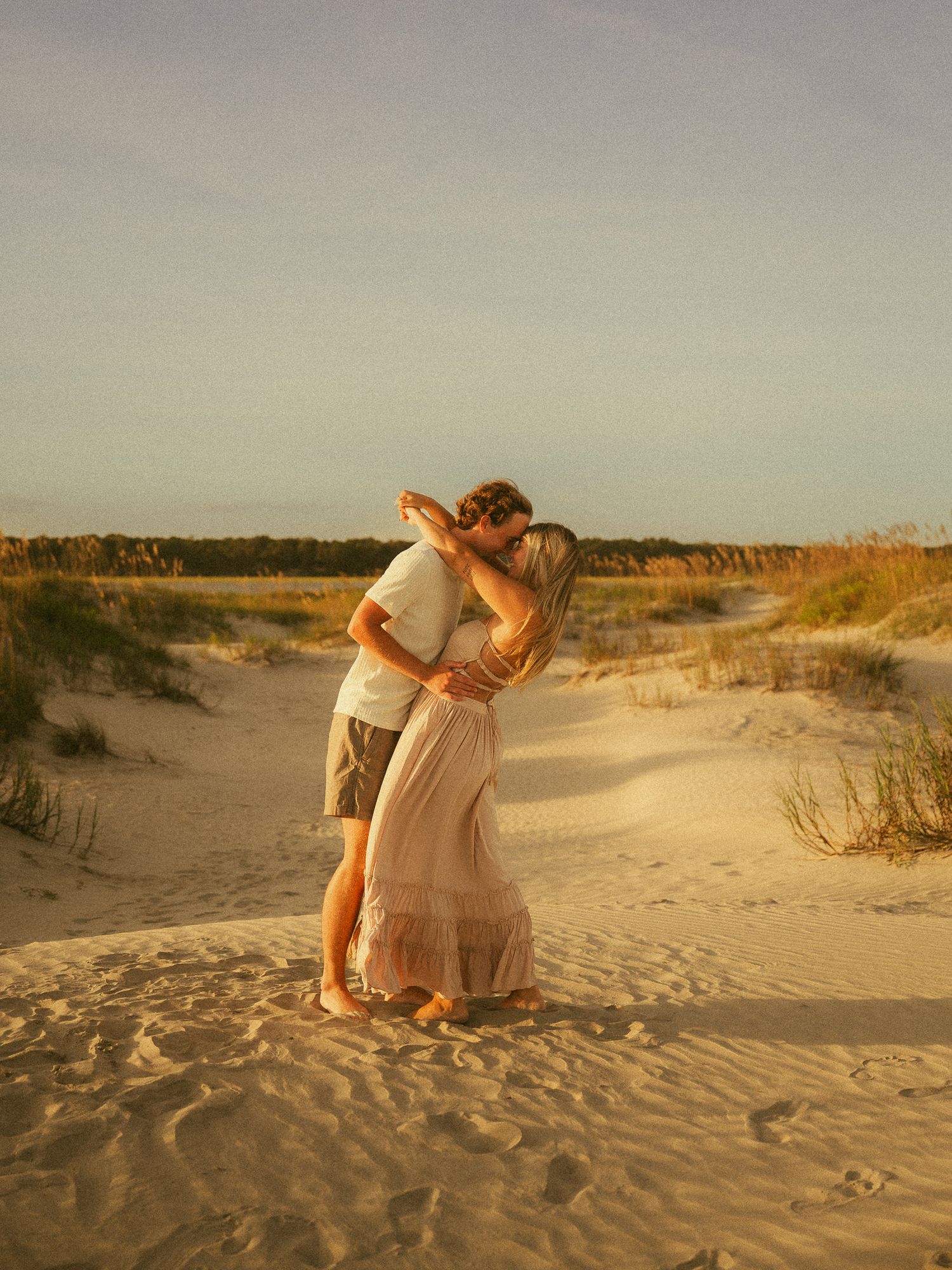 Couple sharing a romantic kiss on sandy beach dunes at golden hour sunset, woman in flowing pink dress.