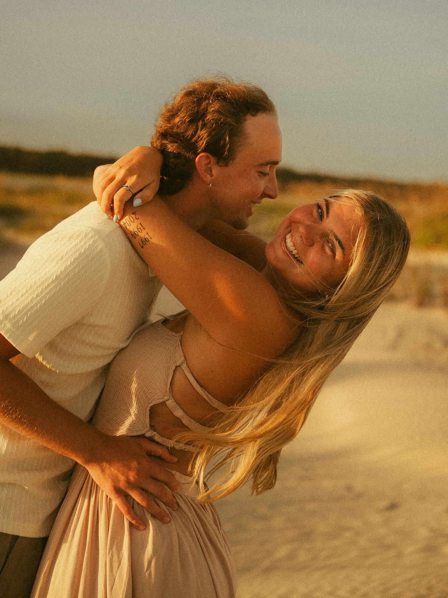 Couple embracing and laughing on a sandy beach at golden hour, woman in pink dress with long blonde hair.