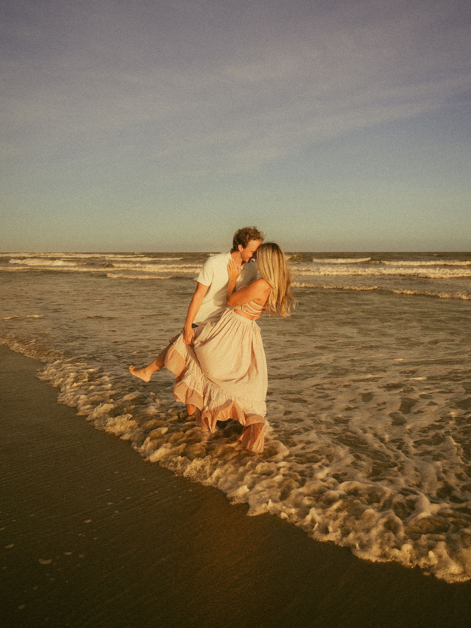 Couple kissing at sunset on a beach, waves washing over their feet, golden hour light, romantic ocean photography.