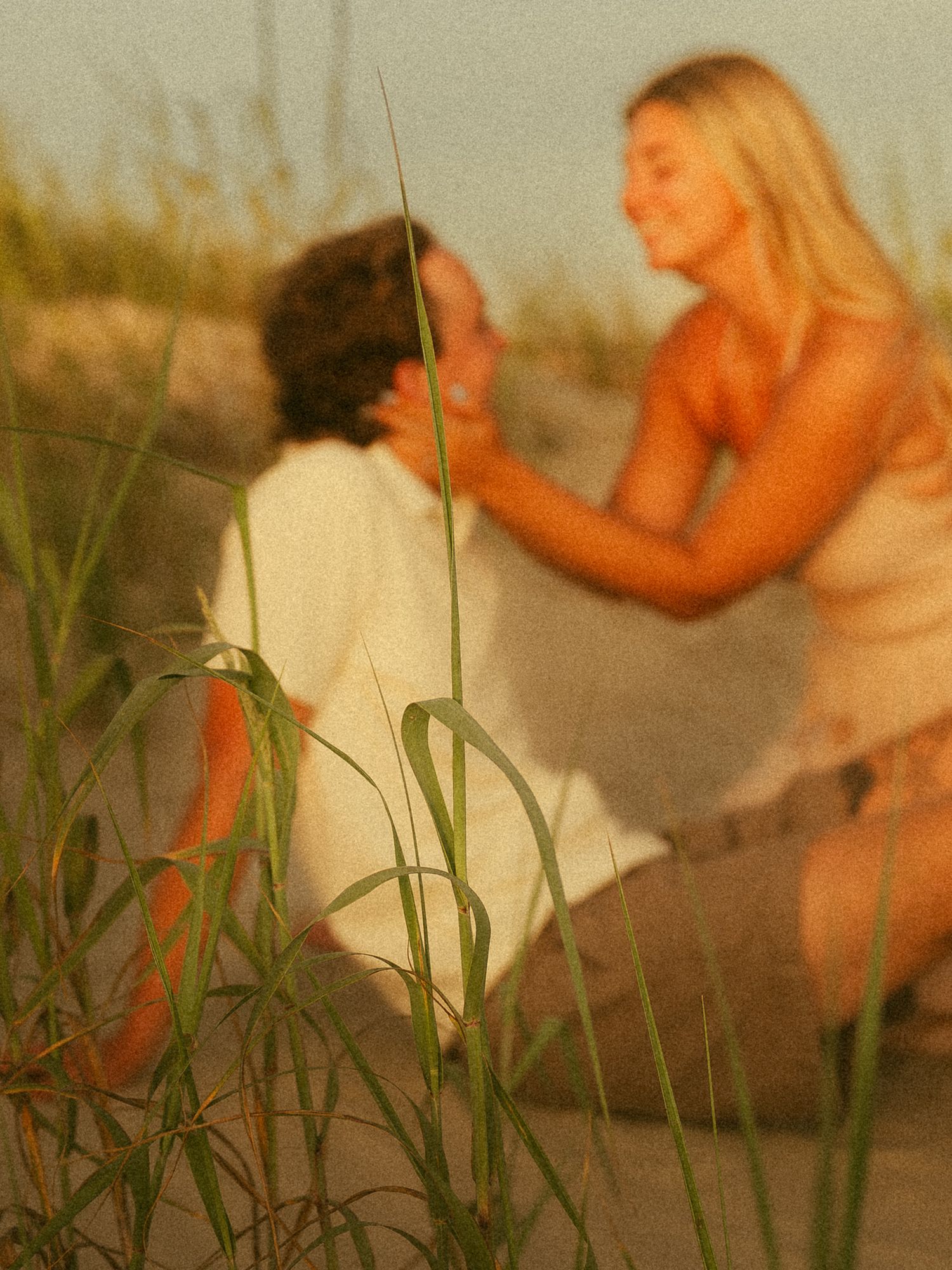 Two people share a tender moment among beach grass dunes at sunset.