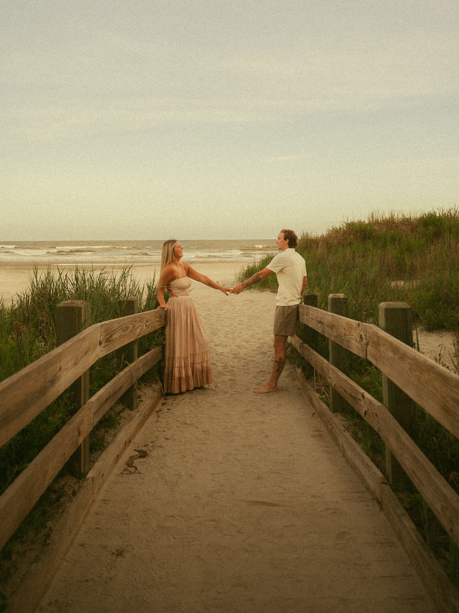 Couple holding hands on a wooden boardwalk leading to a sandy beach at sunset, surrounded by dunes and sea grass.