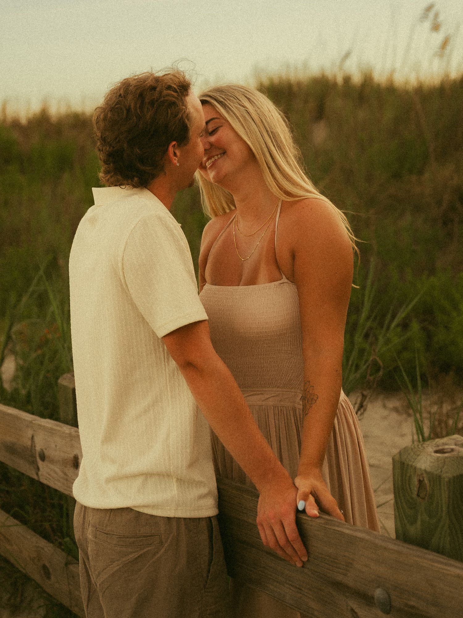A couple shares a romantic moment on a wooden fence at sunset, with coastal dunes and grass in the background.