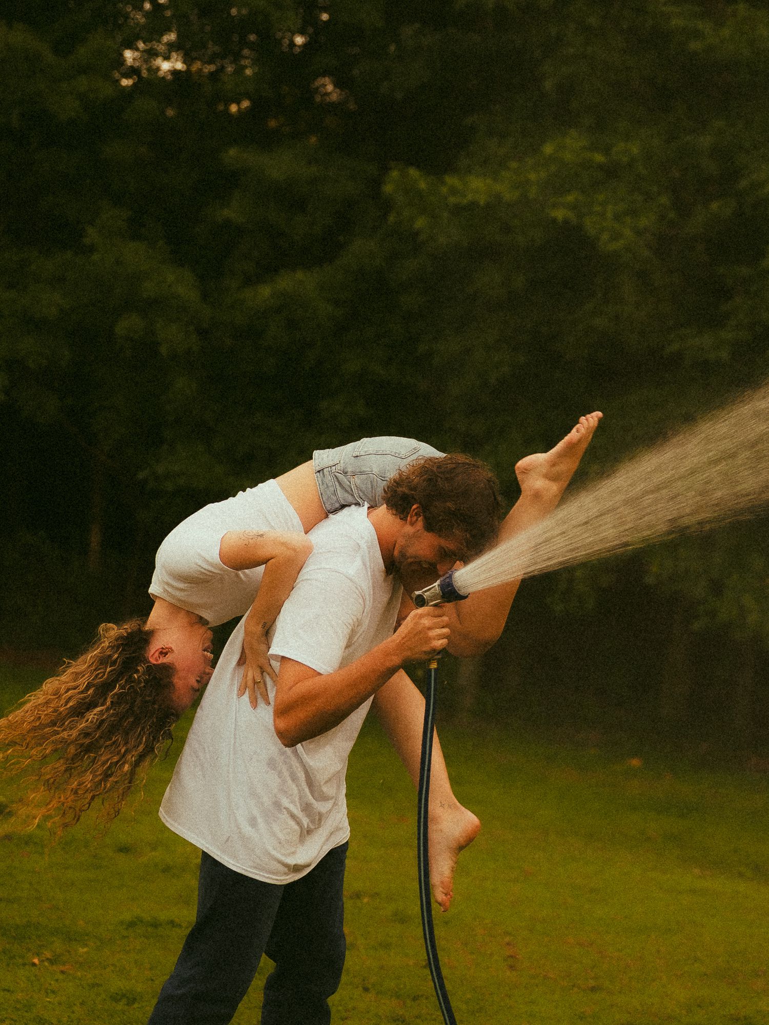 Man in white t-shirt carrying a person over his shoulder while spraying a garden hose outdoors.
