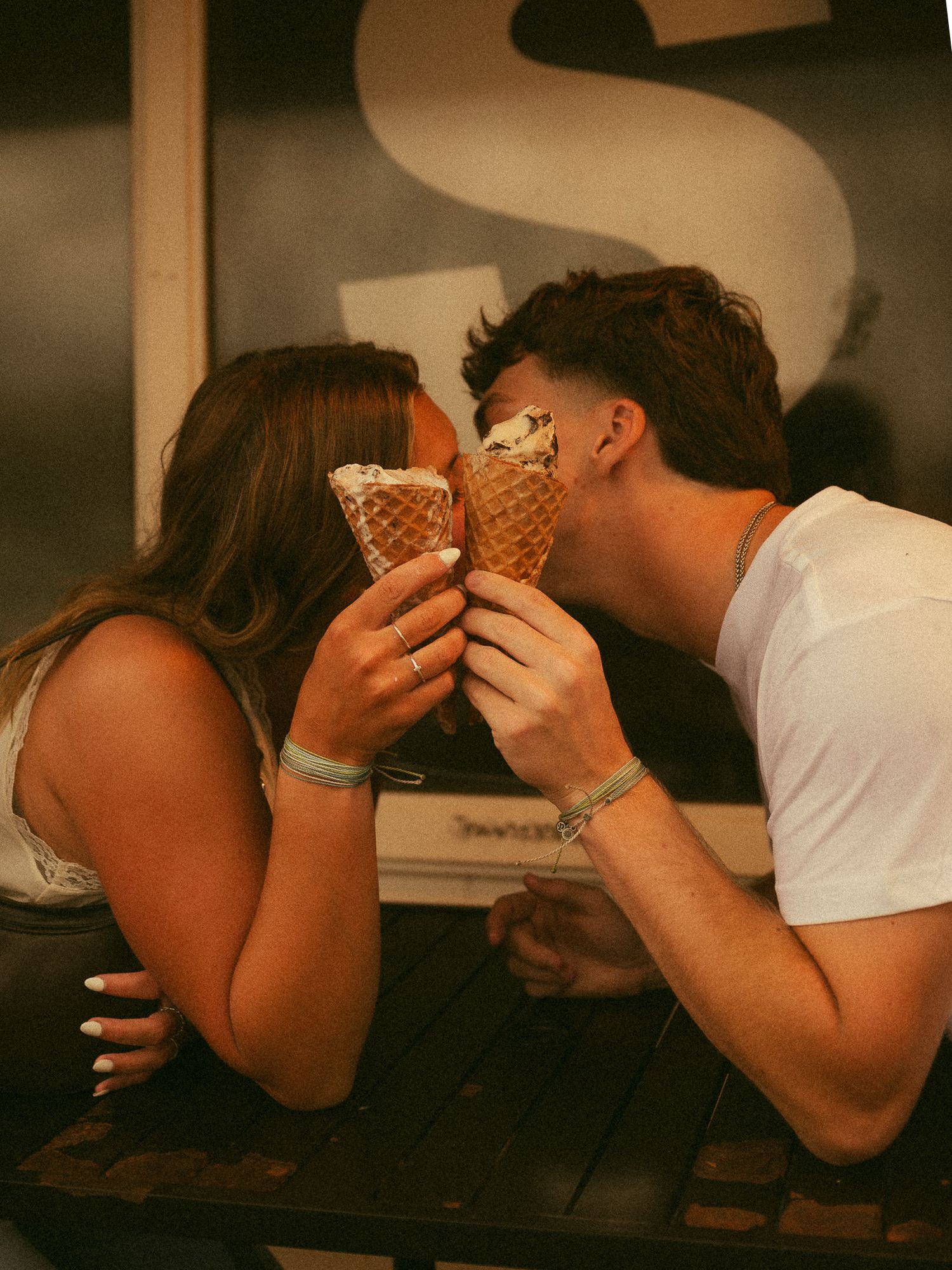 Romantic couple sharing ice cream cones in waffle cones, leaning in for a kiss outdoors.