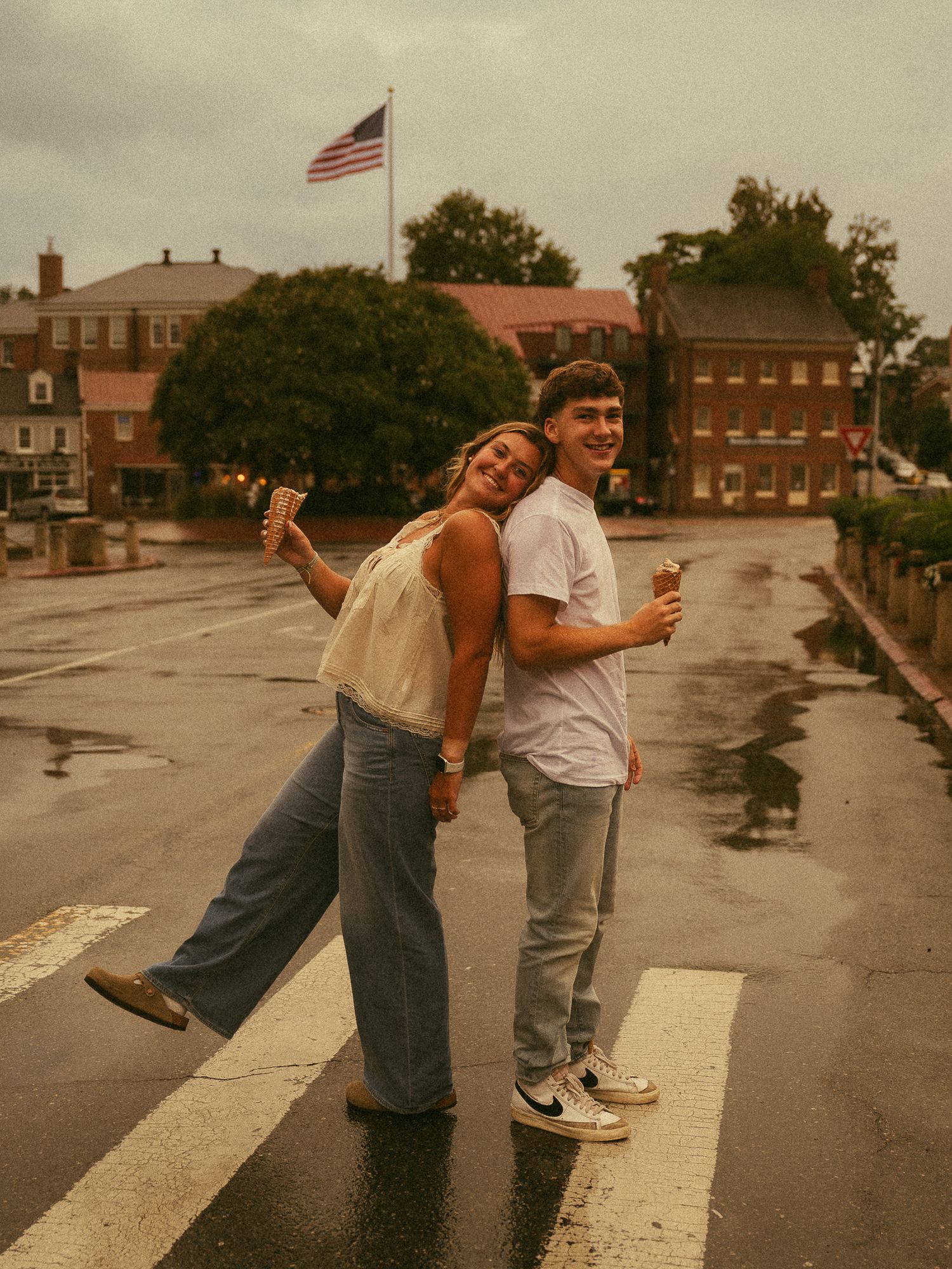 Couple holding ice cream cones posing back-to-back on a wet crosswalk with an American flag in the background.