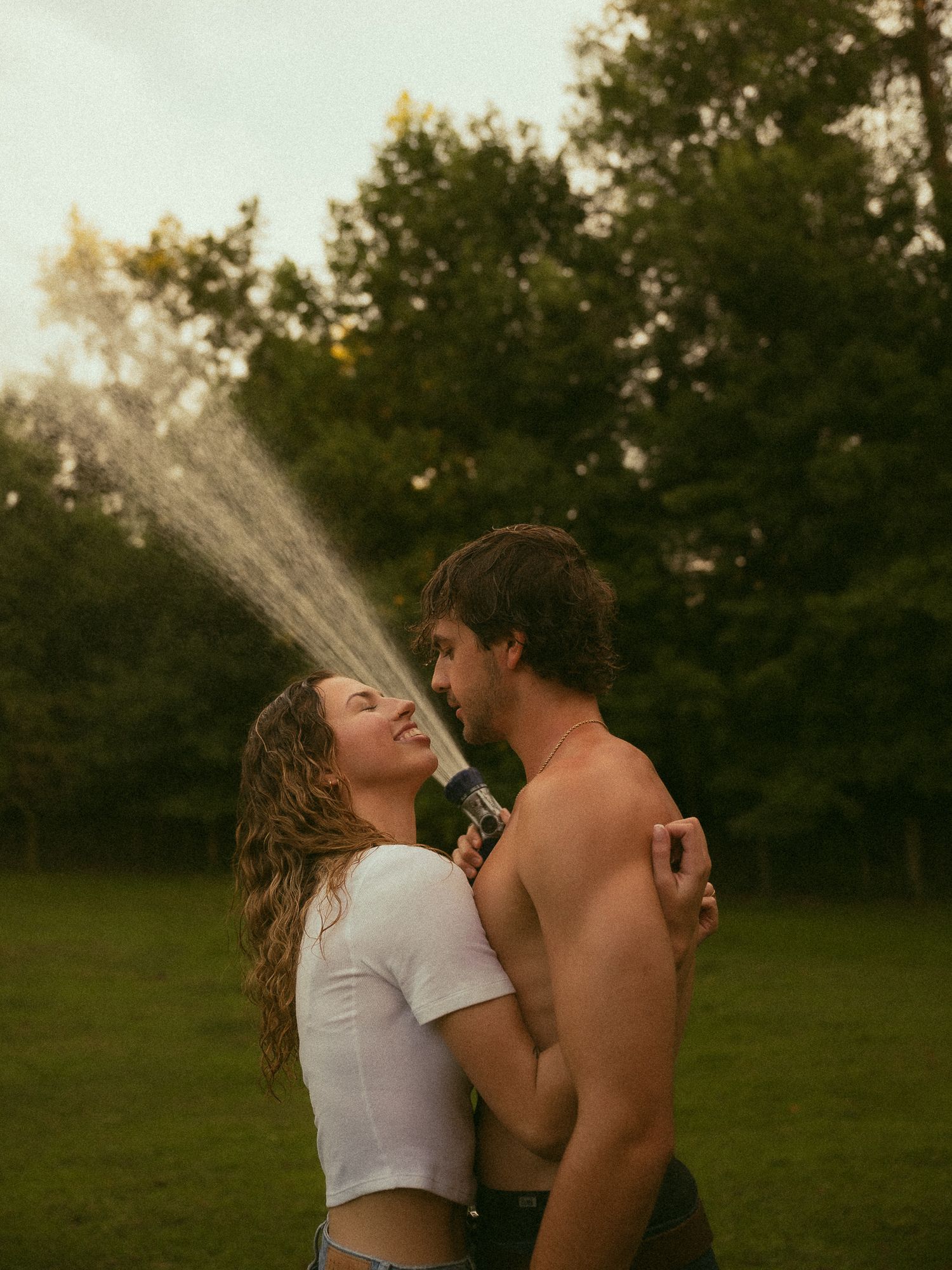 A couple laughs and embraces outdoors while playfully spraying water from a hose in a lush green yard.