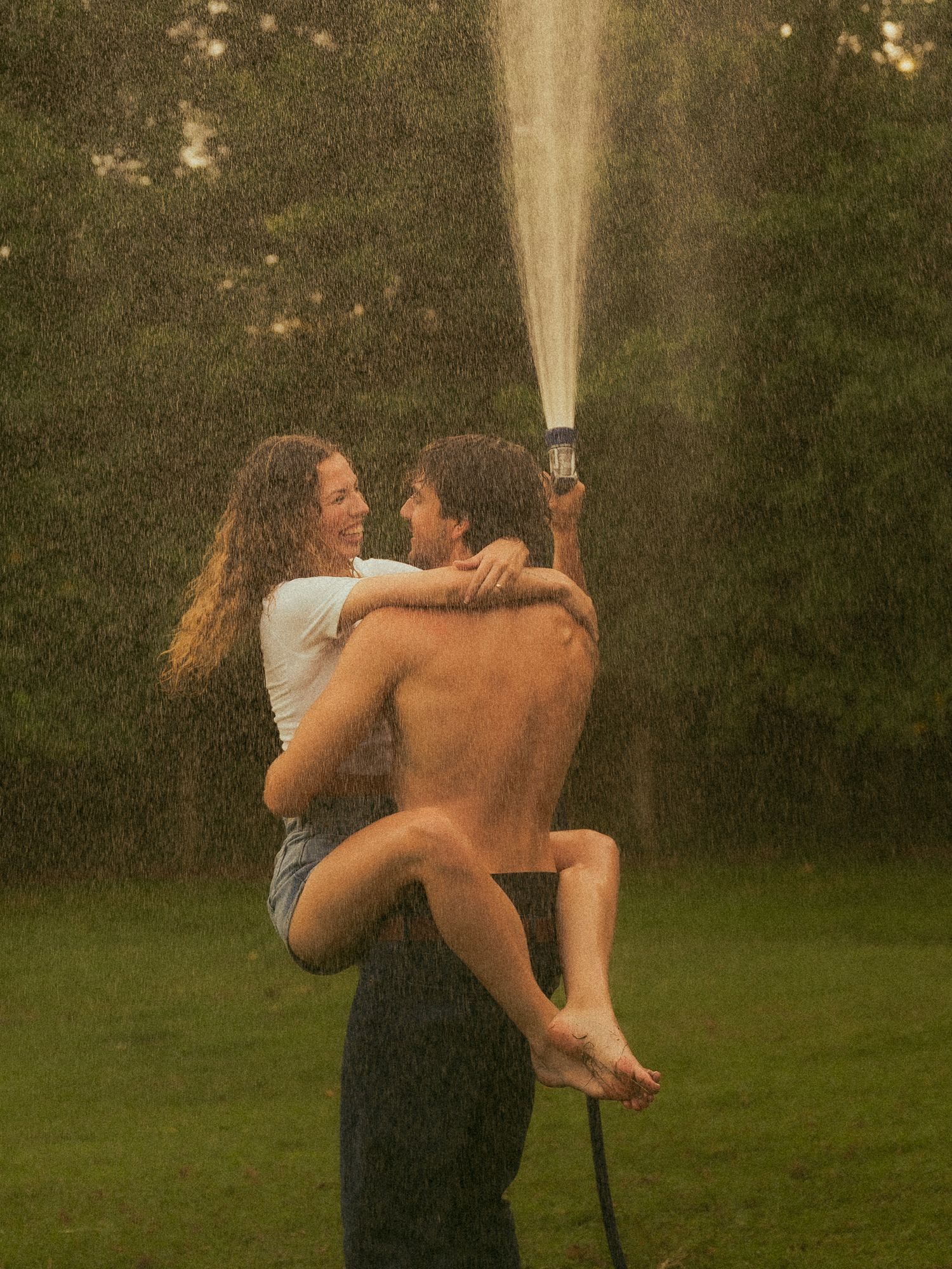 Couple laughing and embracing in backyard sprinkler spray on lush green lawn during summer.
