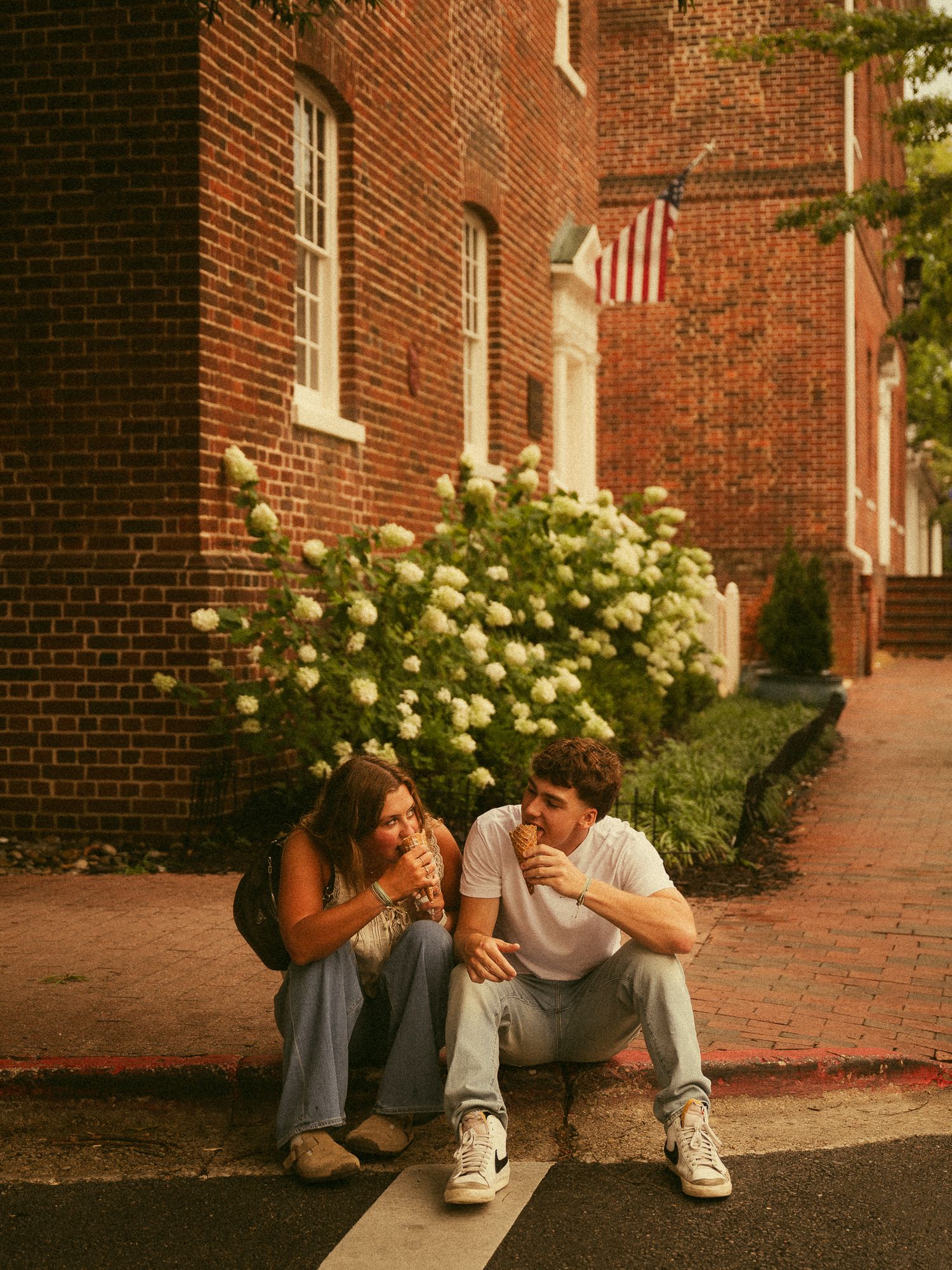 Two young people sitting on a curb eating ice cream beside a brick building with blooming hydrangeas and an American flag.