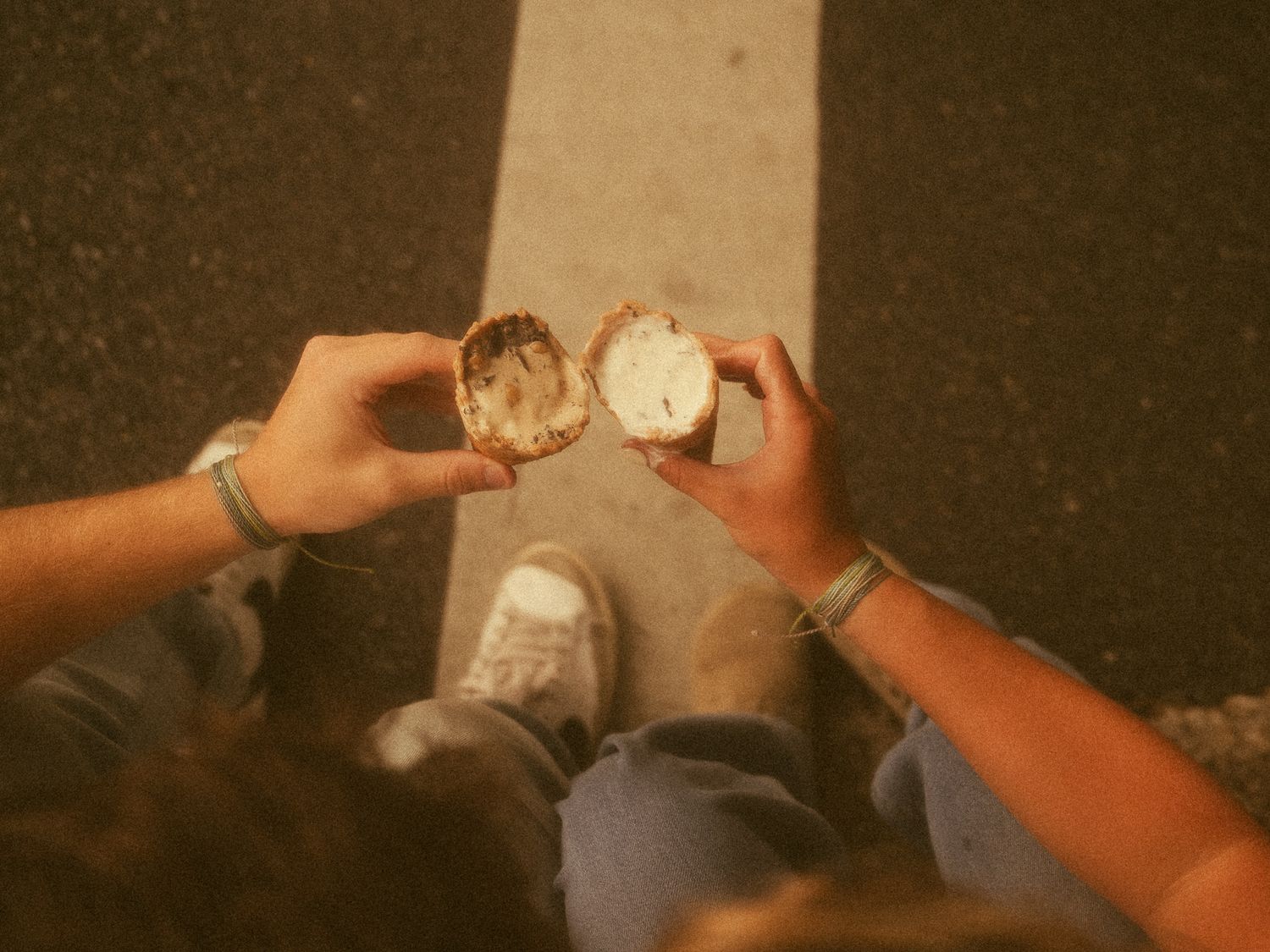 Two people holding a split open coconut over asphalt, wearing bracelets and casual jeans.