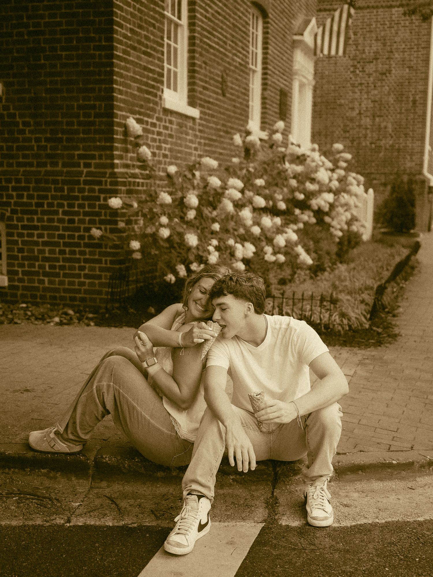 Couple sitting together on a curb in sepia tone, leaning close near a brick building with blooming hydrangeas.