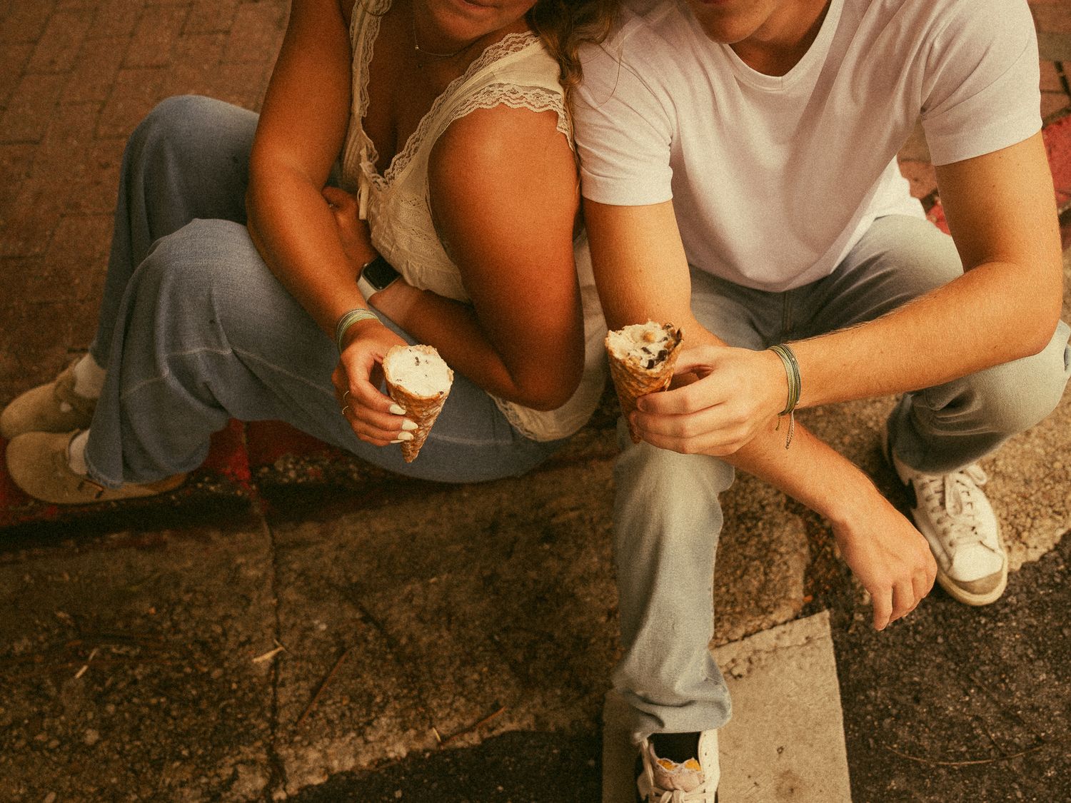 Couple sitting outdoors sharing ice cream cones in waffle cups, wearing casual jeans and sneakers on a brick surface.