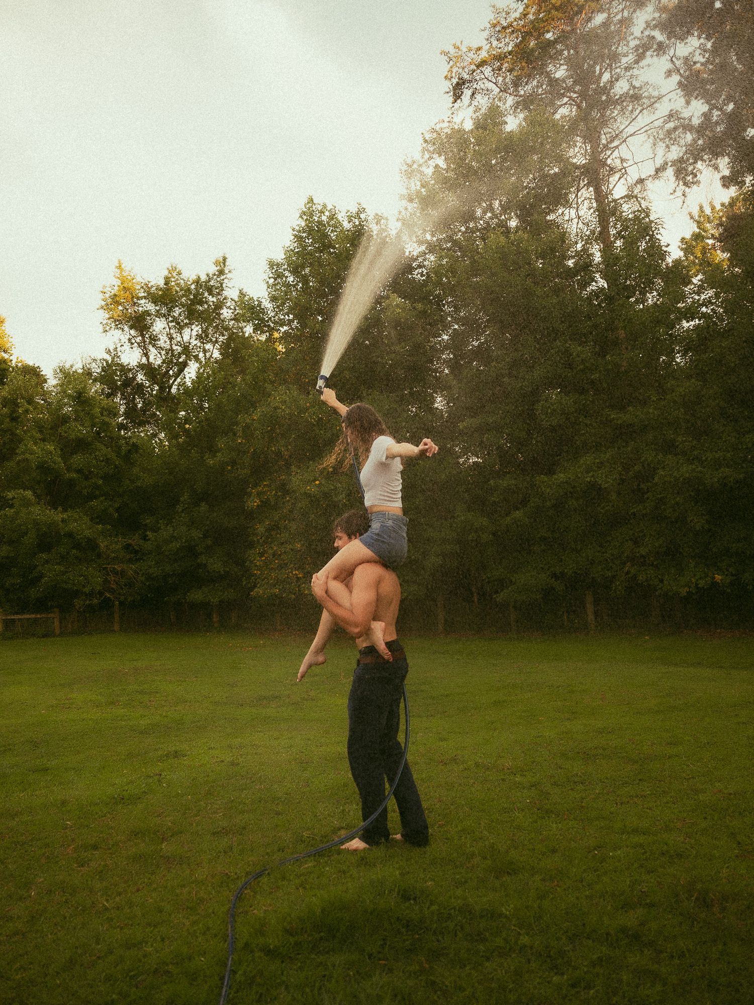 Man lifting woman on shoulders as she sprays water hose in lush green backyard at dusk.