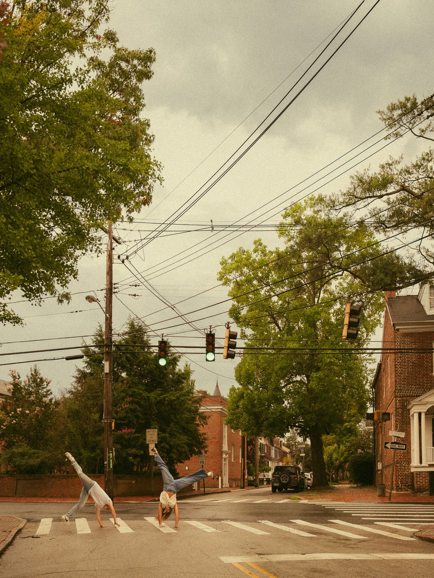 Two people doing cartwheel handstands on a crosswalk in a charming tree-lined brick townhouse neighborhood.
