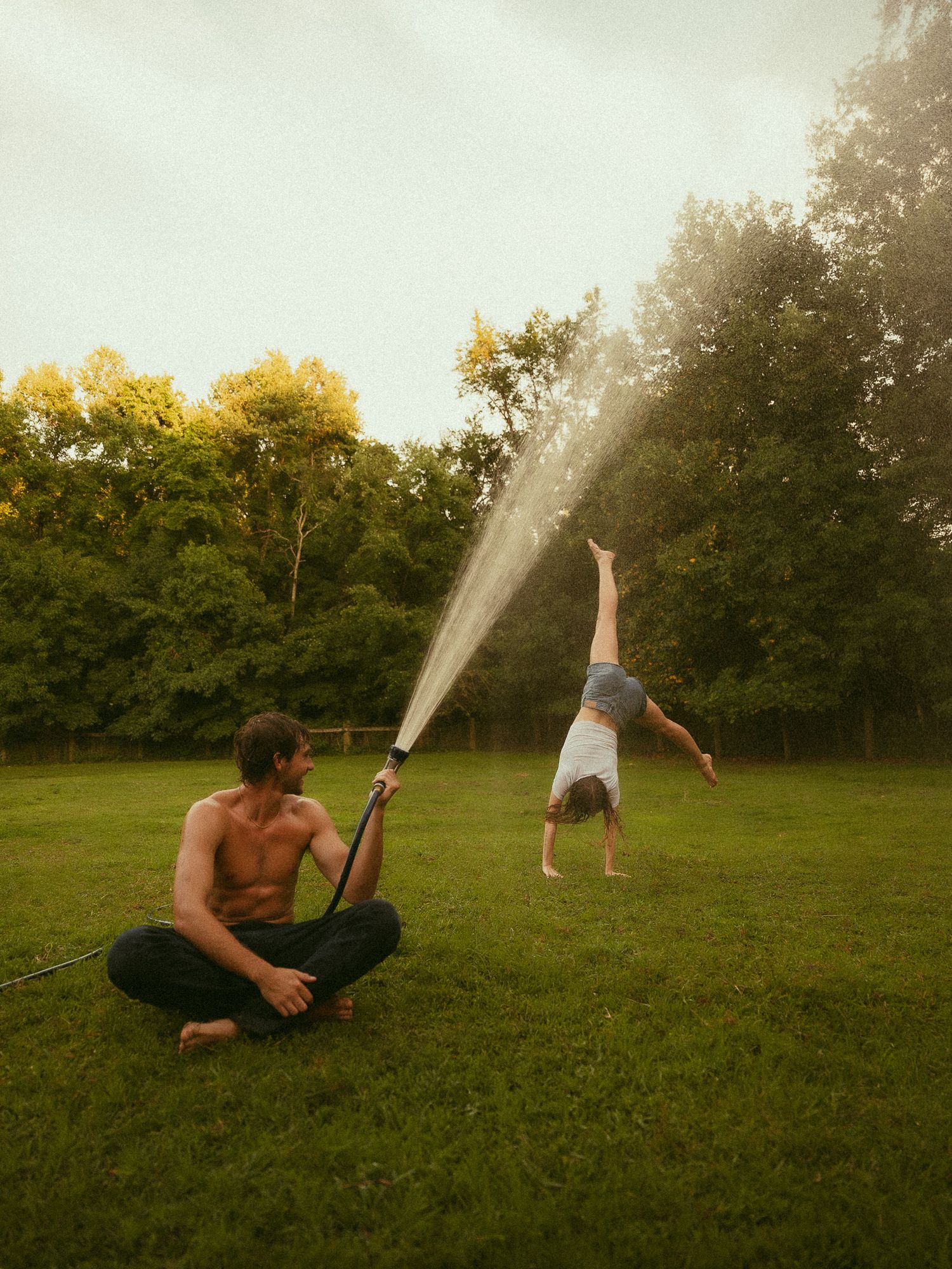 Man sitting cross-legged on grass spraying hose while person does handstand in sunny backyard surrounded by trees.