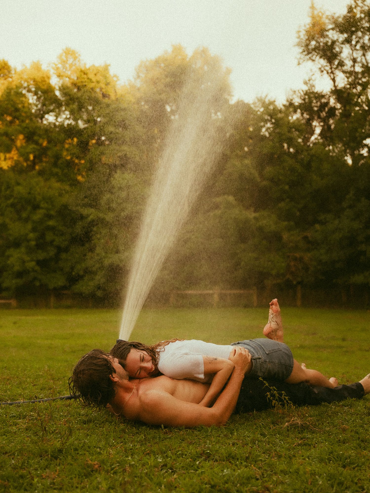 Couple lying on green grass embracing romantically while a lawn sprinkler sprays water above them outdoors.