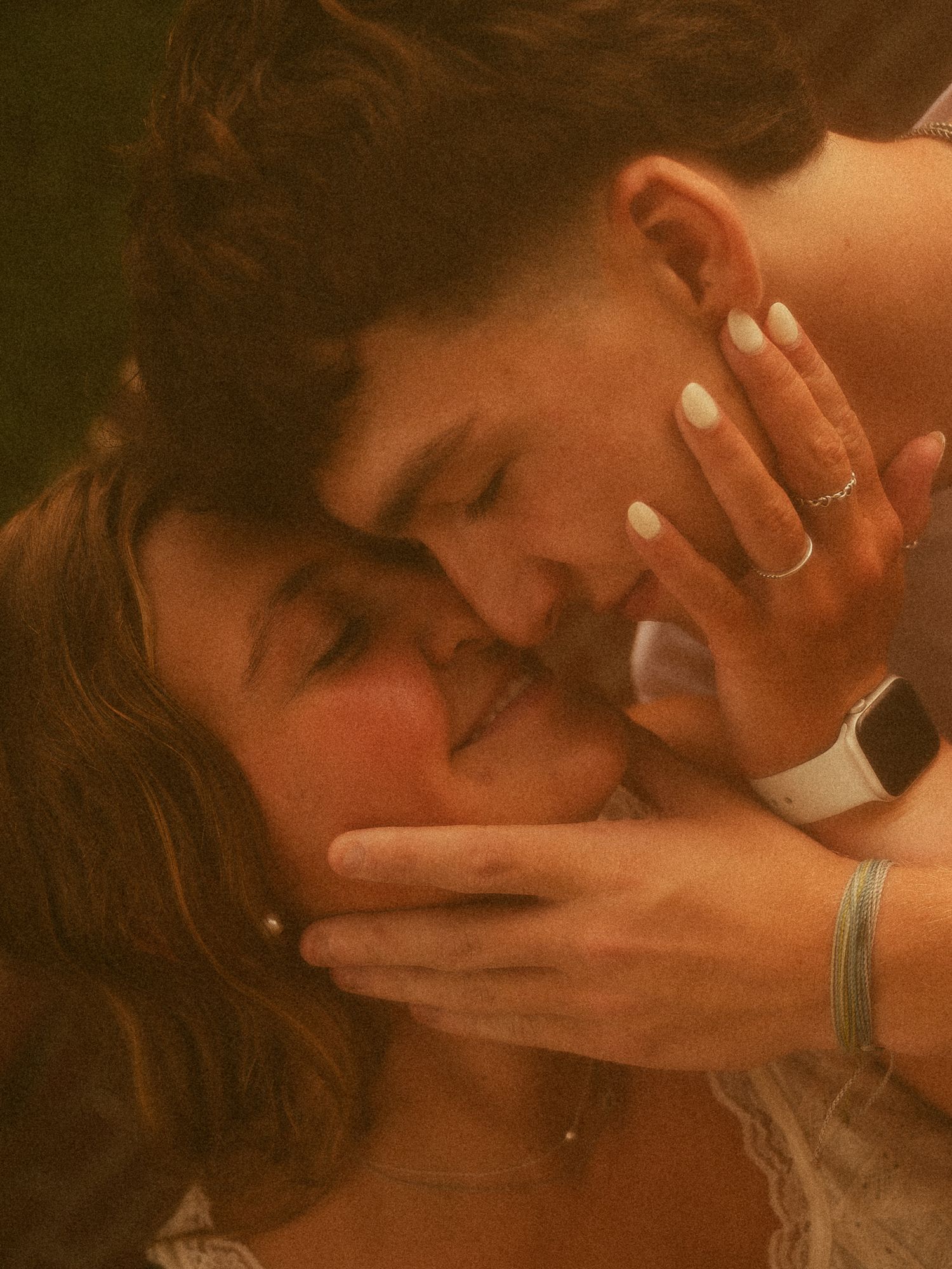 A romantic couple sharing an intimate moment, with hands gently cradling faces, rings and bracelets visible.