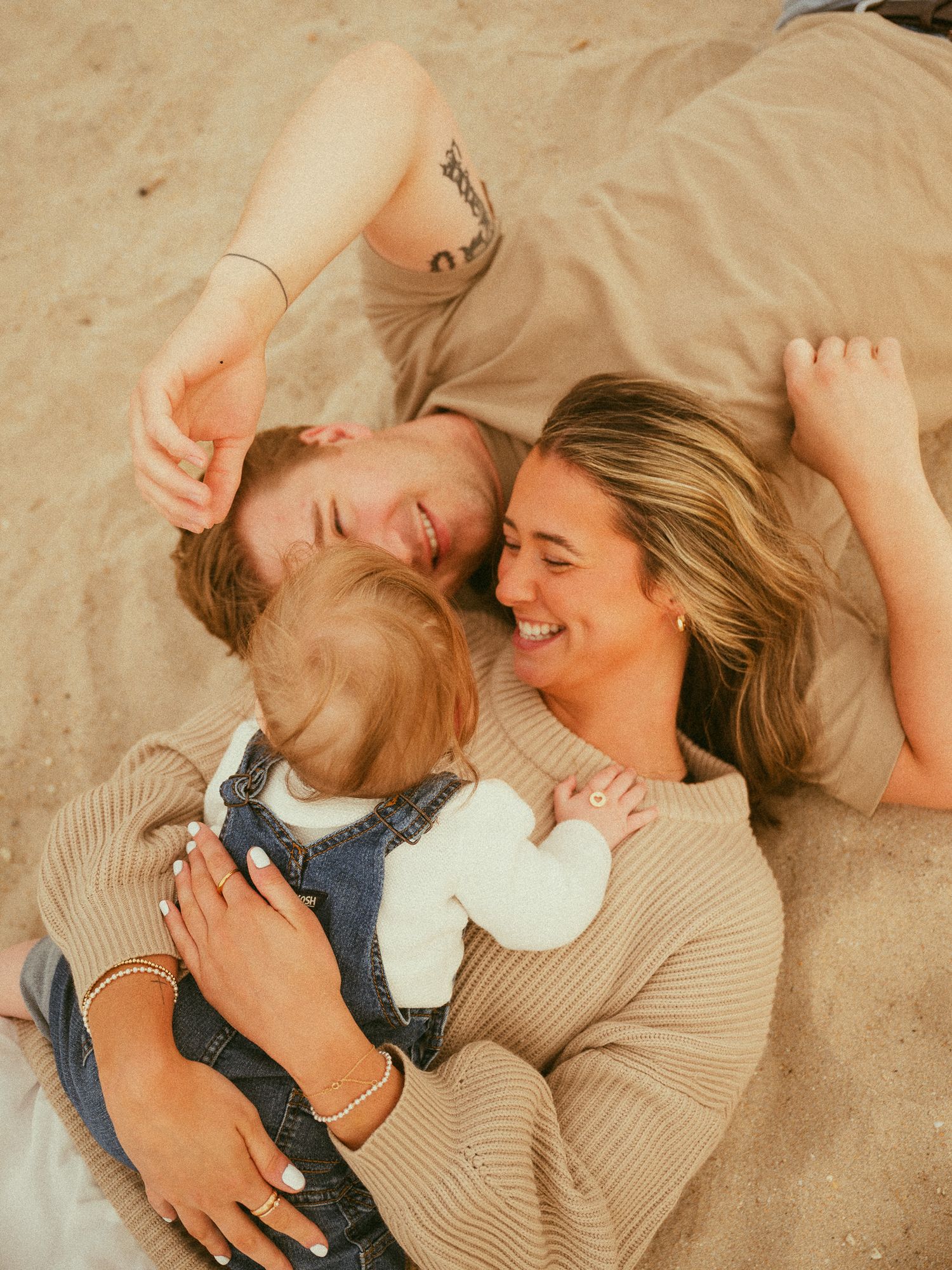 Young couple lying on sandy beach smiling with their baby in a warm, candid family portrait.