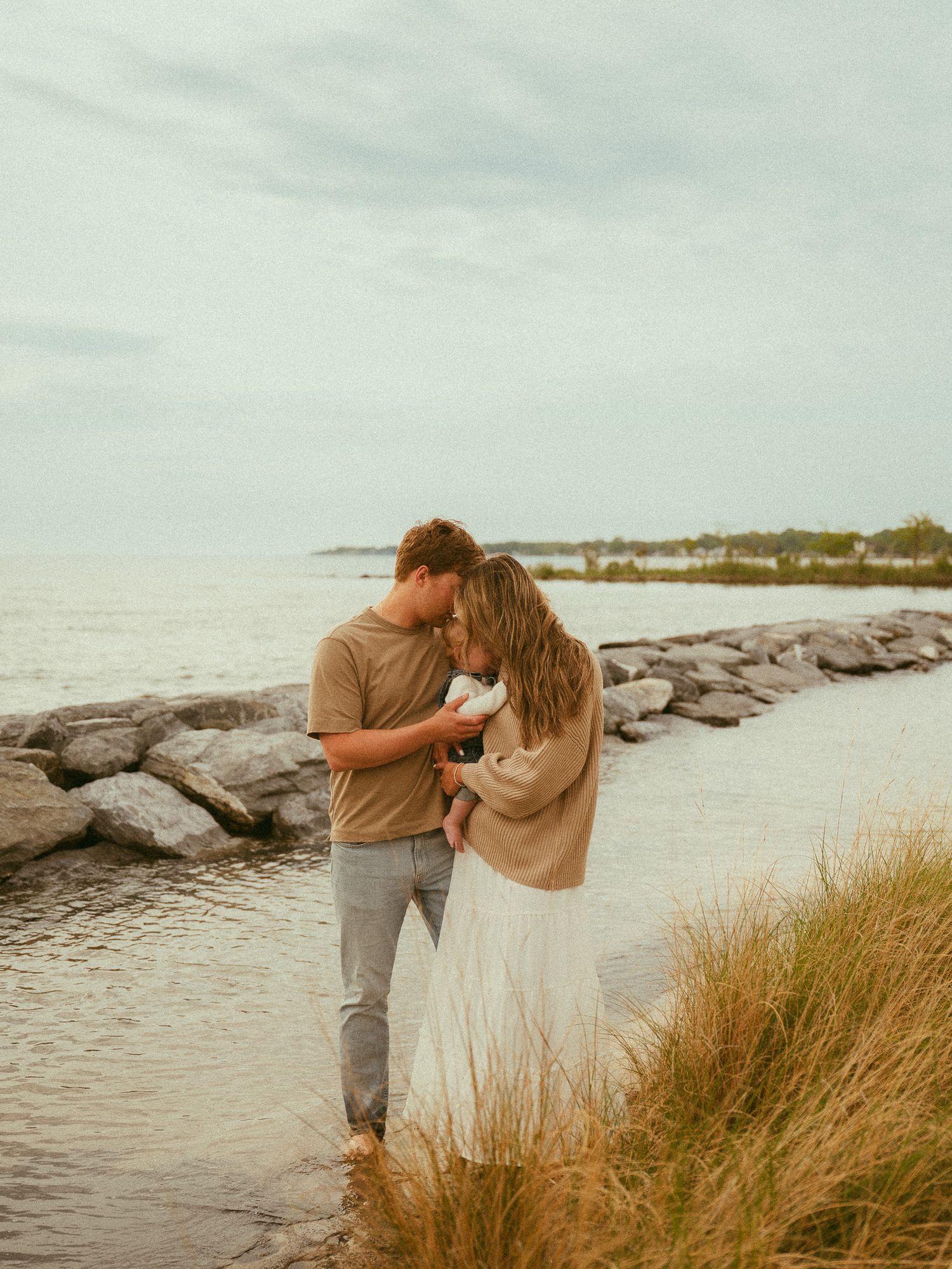 Romantic couple embracing near rocky shoreline with tall grass and calm water in background.