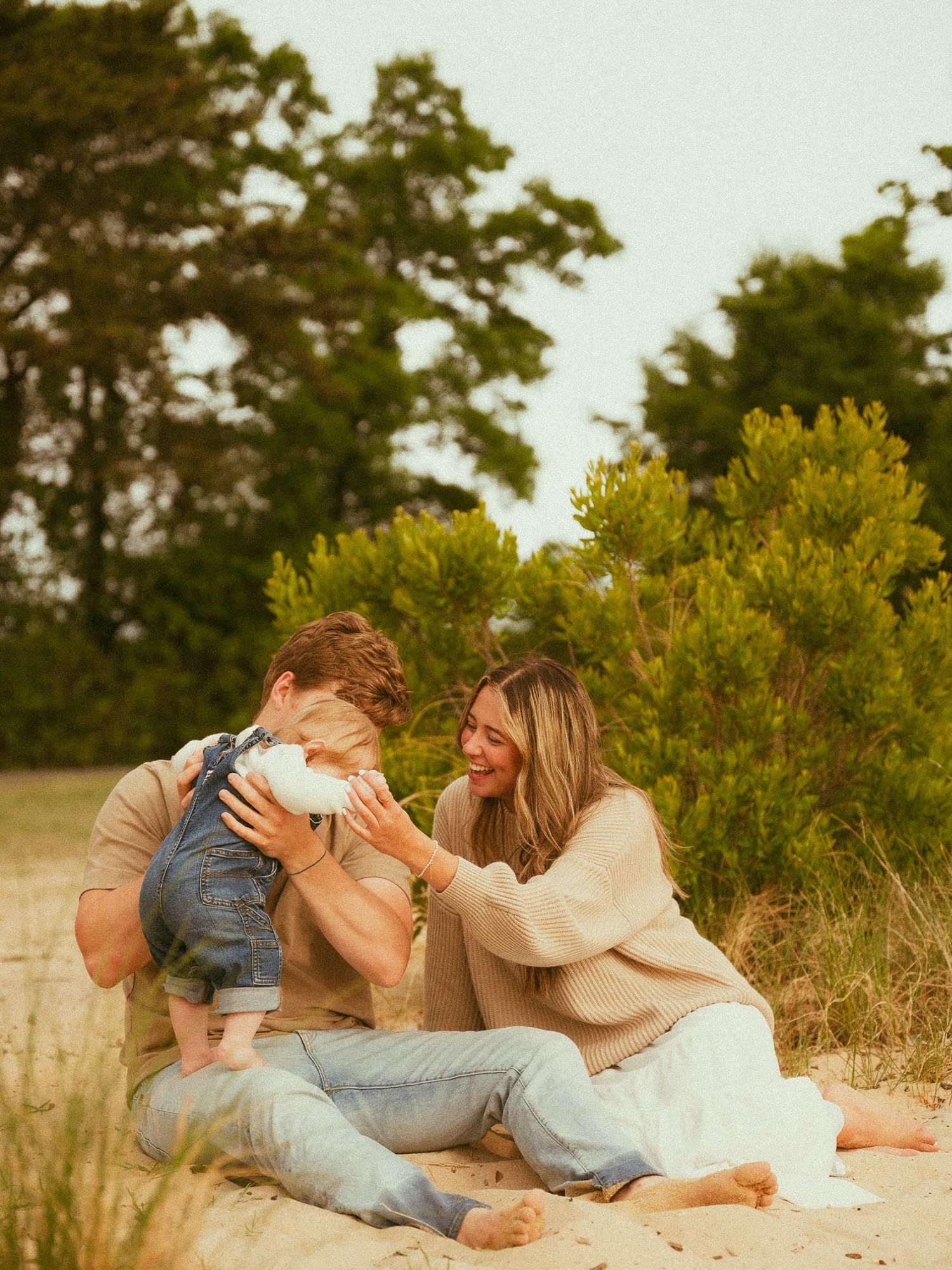 A young family sits together on sandy ground outdoors, laughing and playing with their baby near lush green trees.