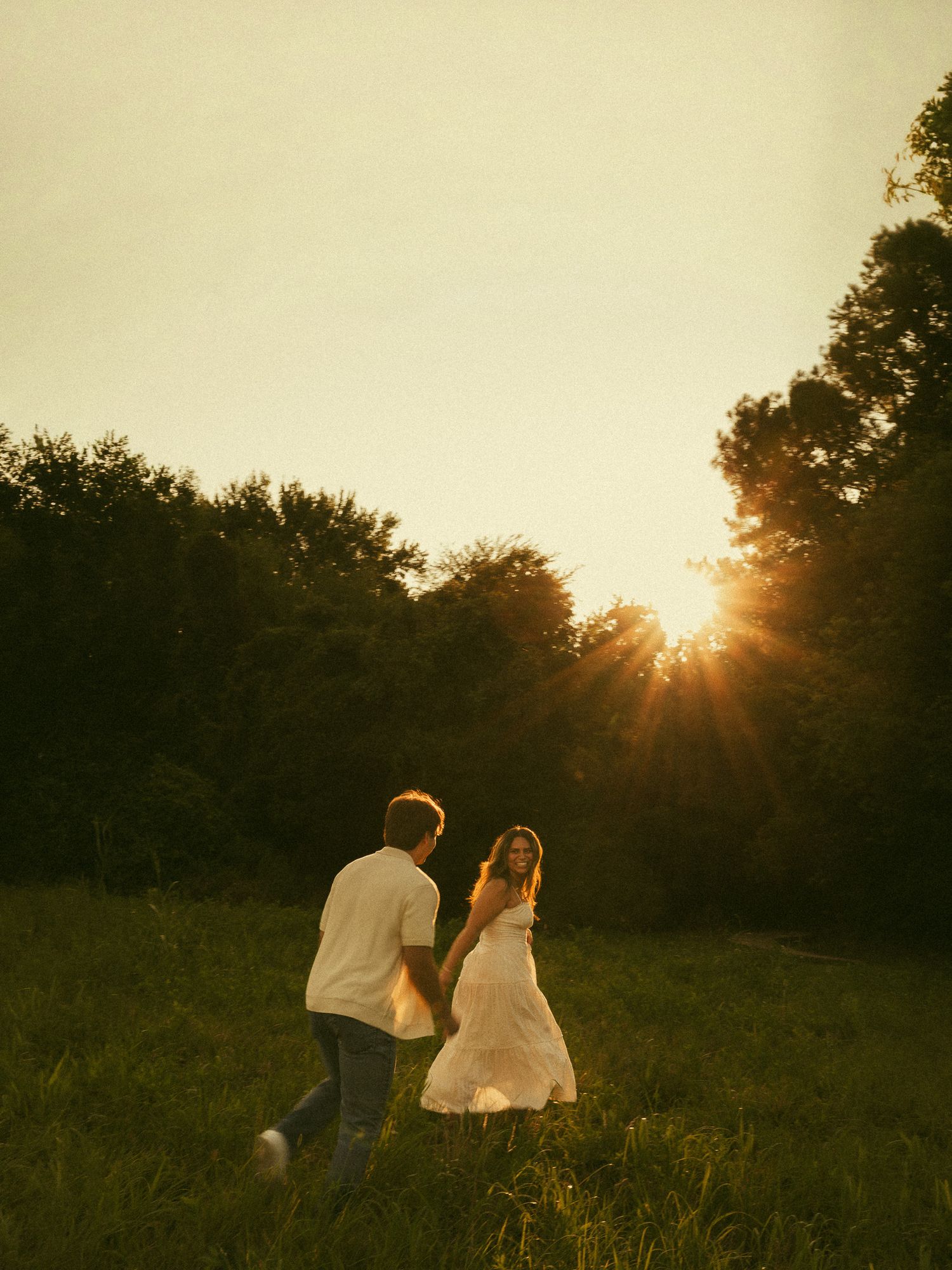 Couple holding hands running through a sunlit green meadow at golden hour with trees and sun rays in the background.