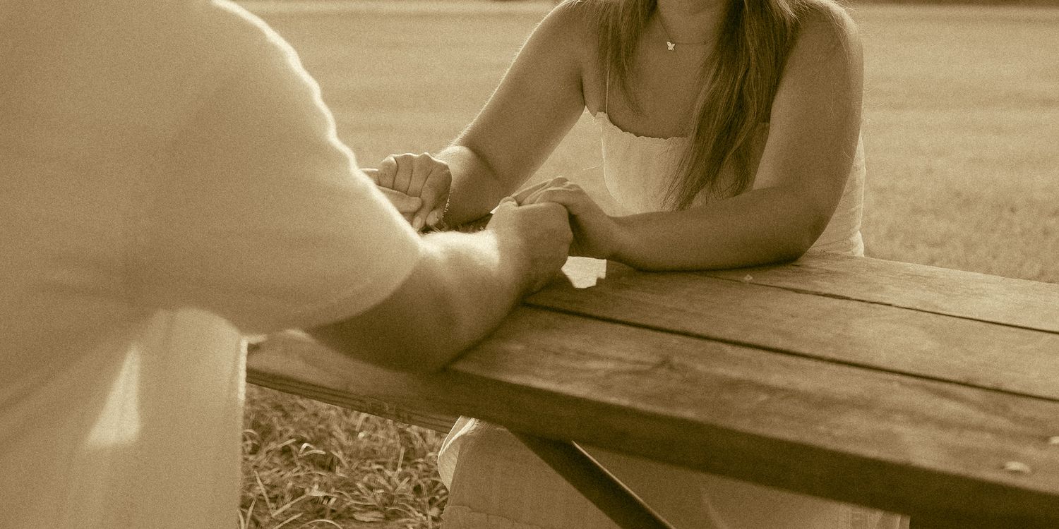 Couple holding hands across a wooden picnic table outdoors in a warm sepia-toned romantic moment.
