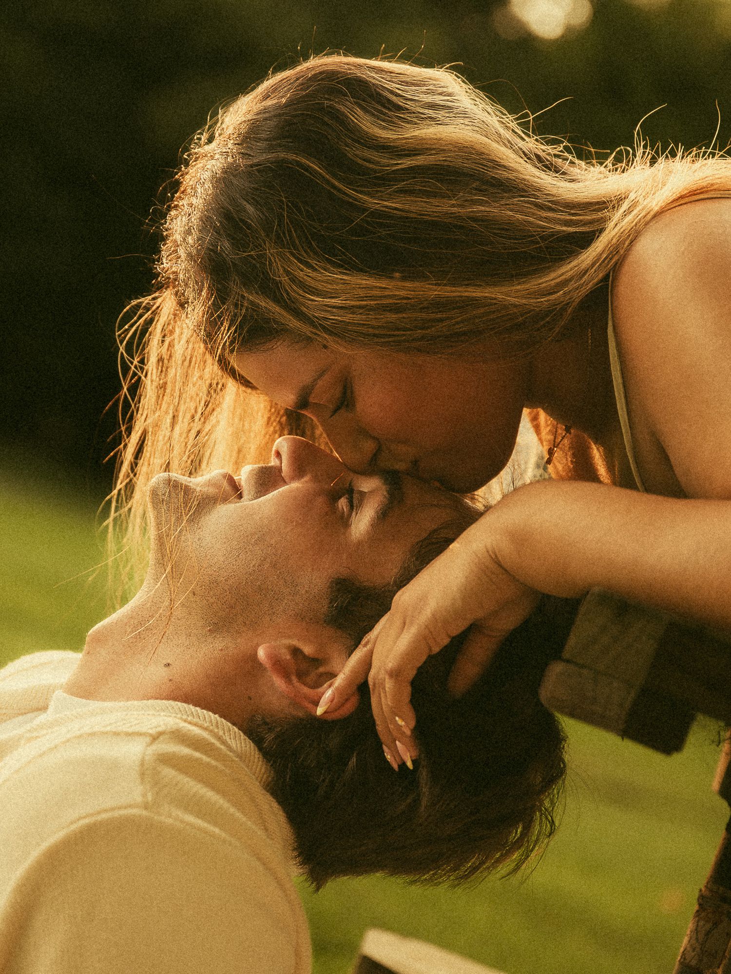A couple shares a tender kiss outdoors in golden sunlight, with the woman leaning over the man lying down.