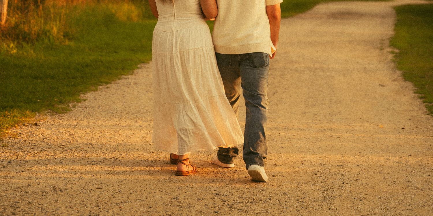 Couple walking hand in hand down a sunlit gravel path at sunset, woman in white dress and man in jeans.
