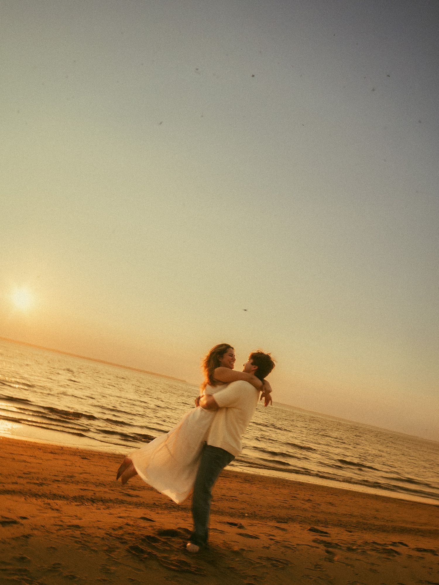 Couple embracing on a sandy beach at golden sunset, man lifting woman in white dress by the ocean shore.