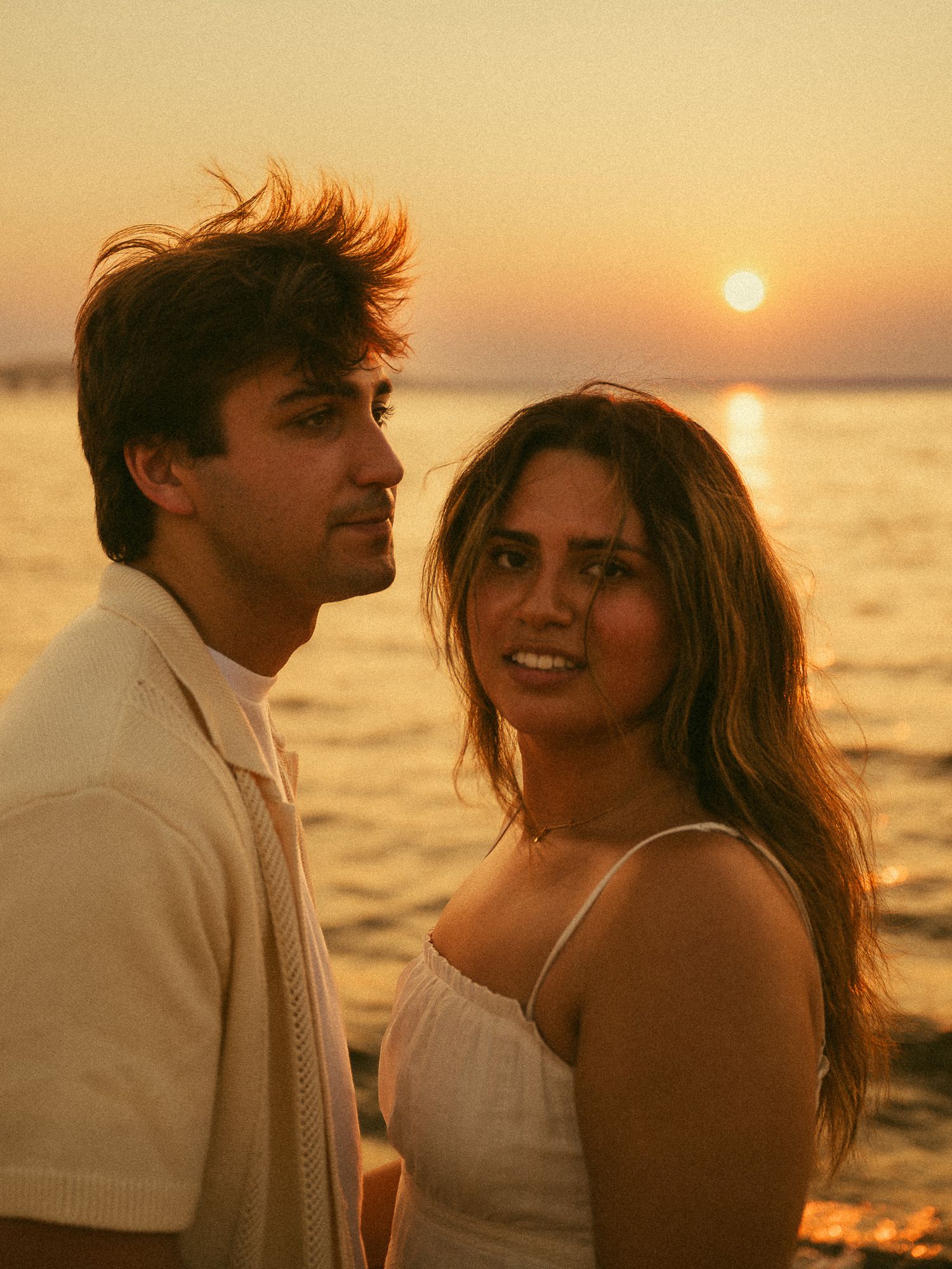 Couple posing romantically by the ocean at golden sunset, woman in white dress smiling at camera.