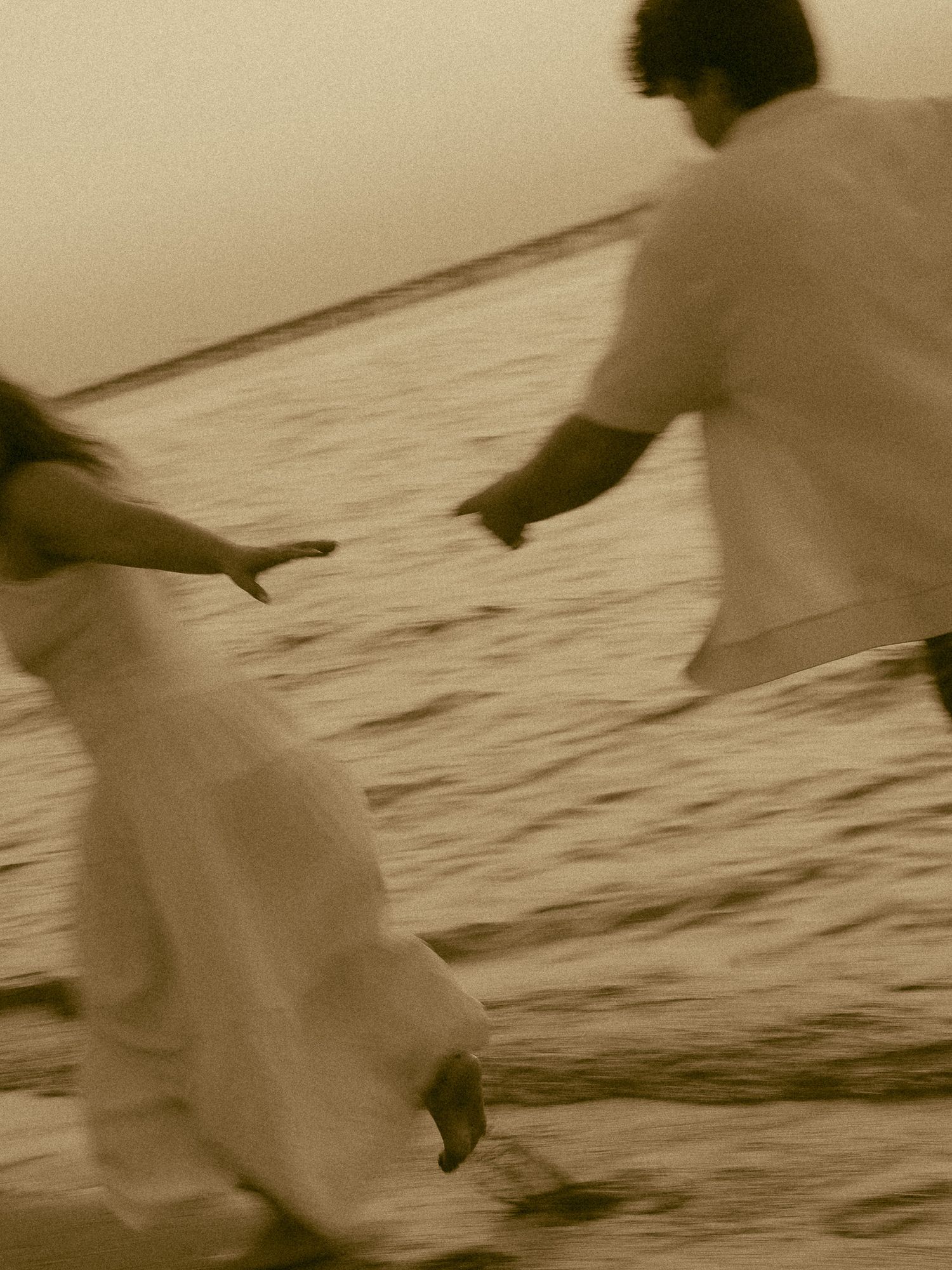 Couple in white clothing running along a beach at sunset in a romantic sepia-toned blurred motion photograph.