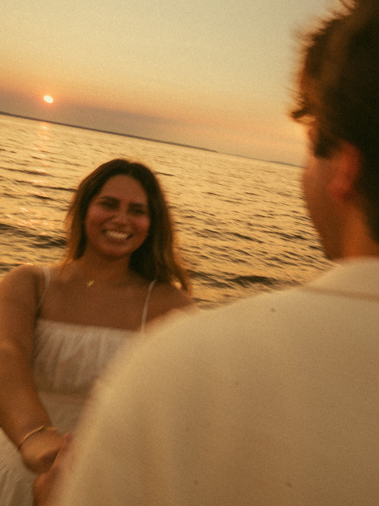 A smiling woman in a white dress holds hands with a man at sunset over the ocean, golden light reflecting on calm waters.