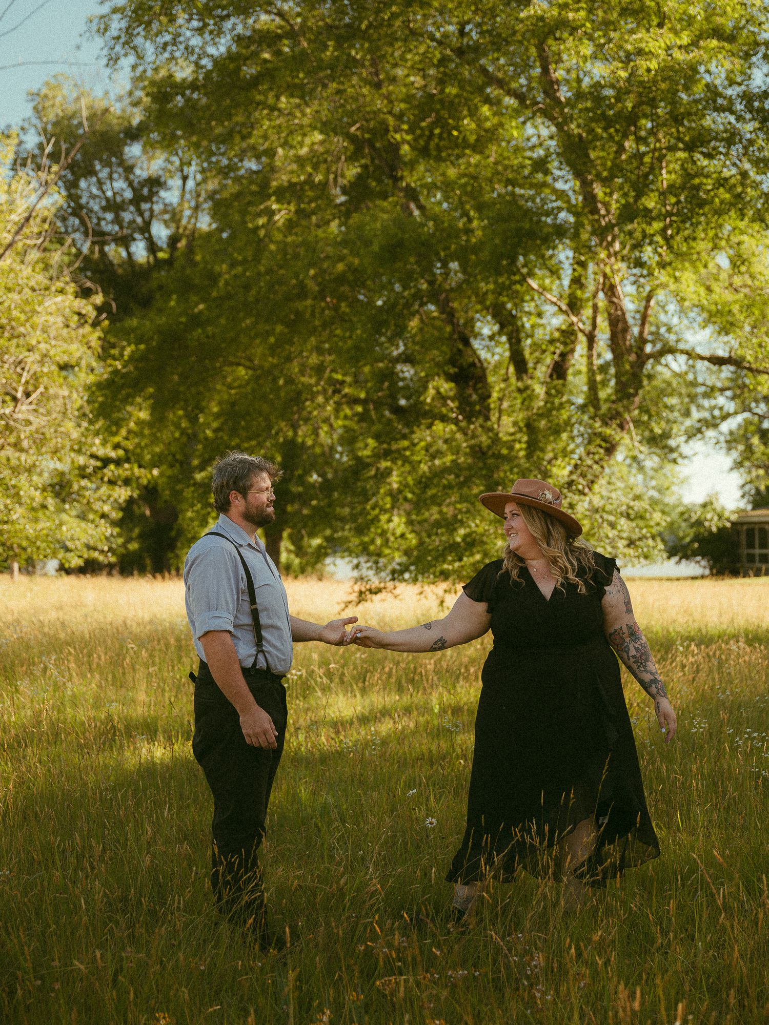 A couple holds hands in a sunlit meadow, the woman in a black dress and cowboy hat, the man in suspenders, with lush trees behind.