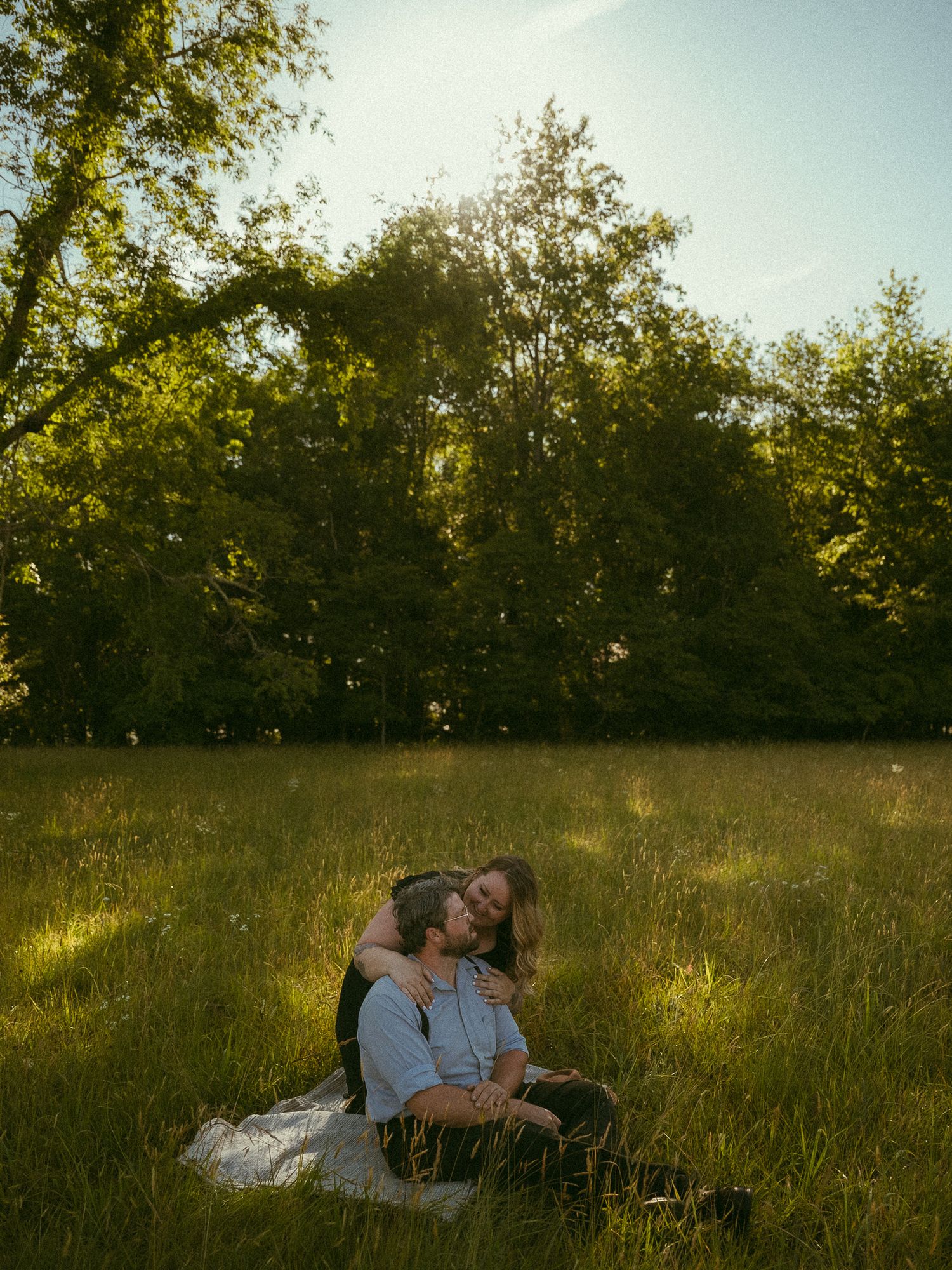 A couple sits together in a sunlit meadow surrounded by lush green trees during golden hour.