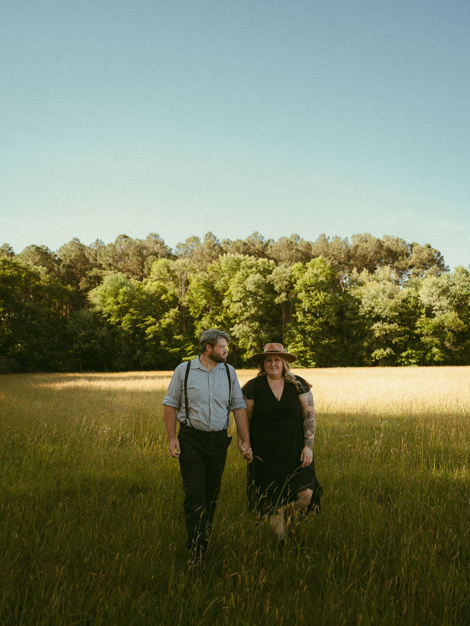 Couple walking hand in hand through a sunny green meadow with lush trees in the background on a clear day.
