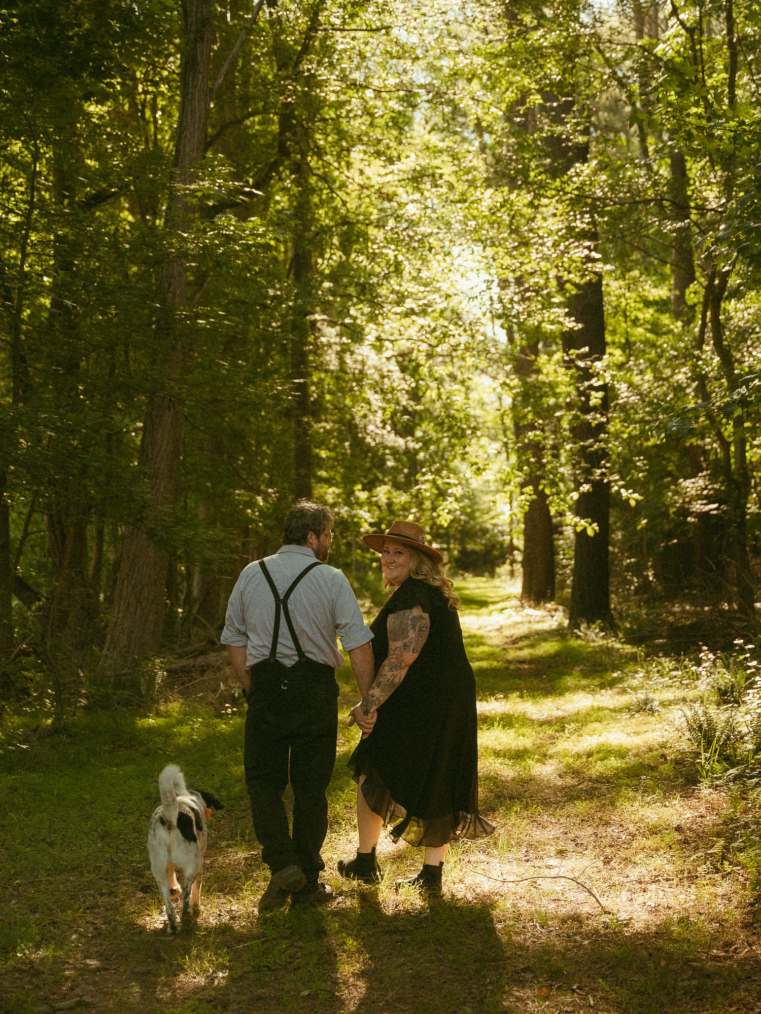 Couple walking a white dog on a sunlit forest path, holding hands, wearing black and white outfits.